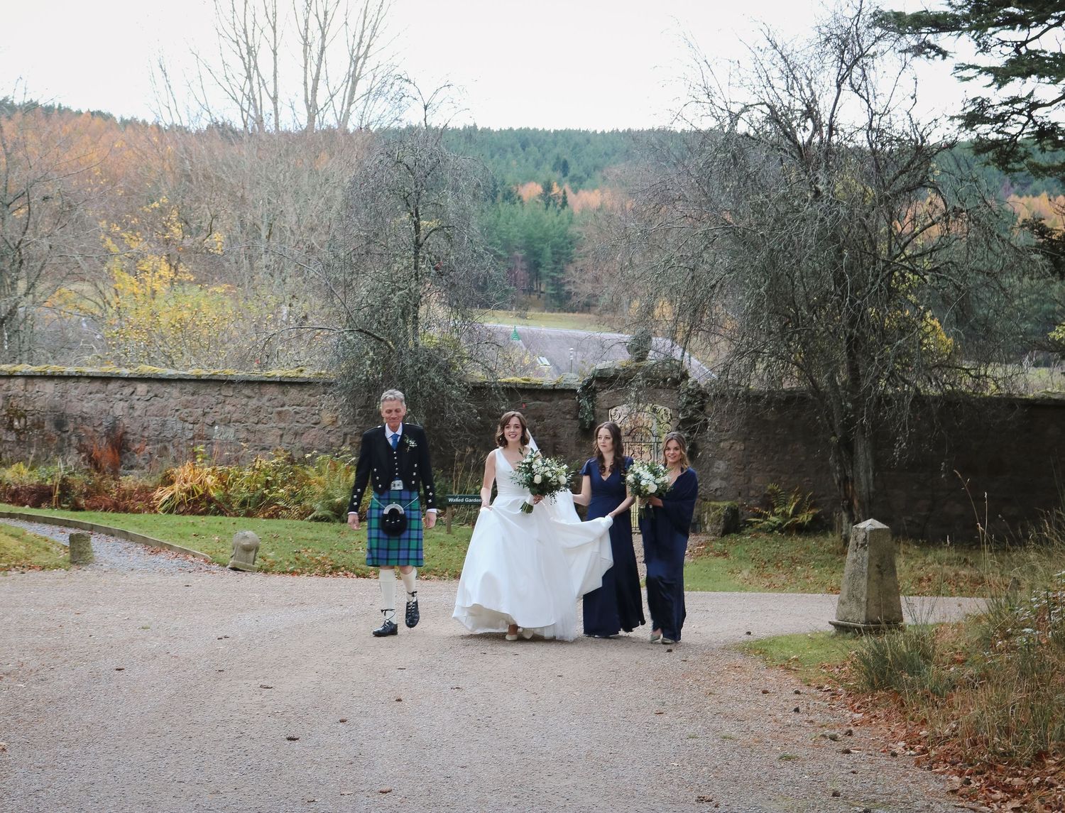A scenic autumn wedding party walks along a gravel path beside a tranquil lake surrounded by bare trees and rolling hills.