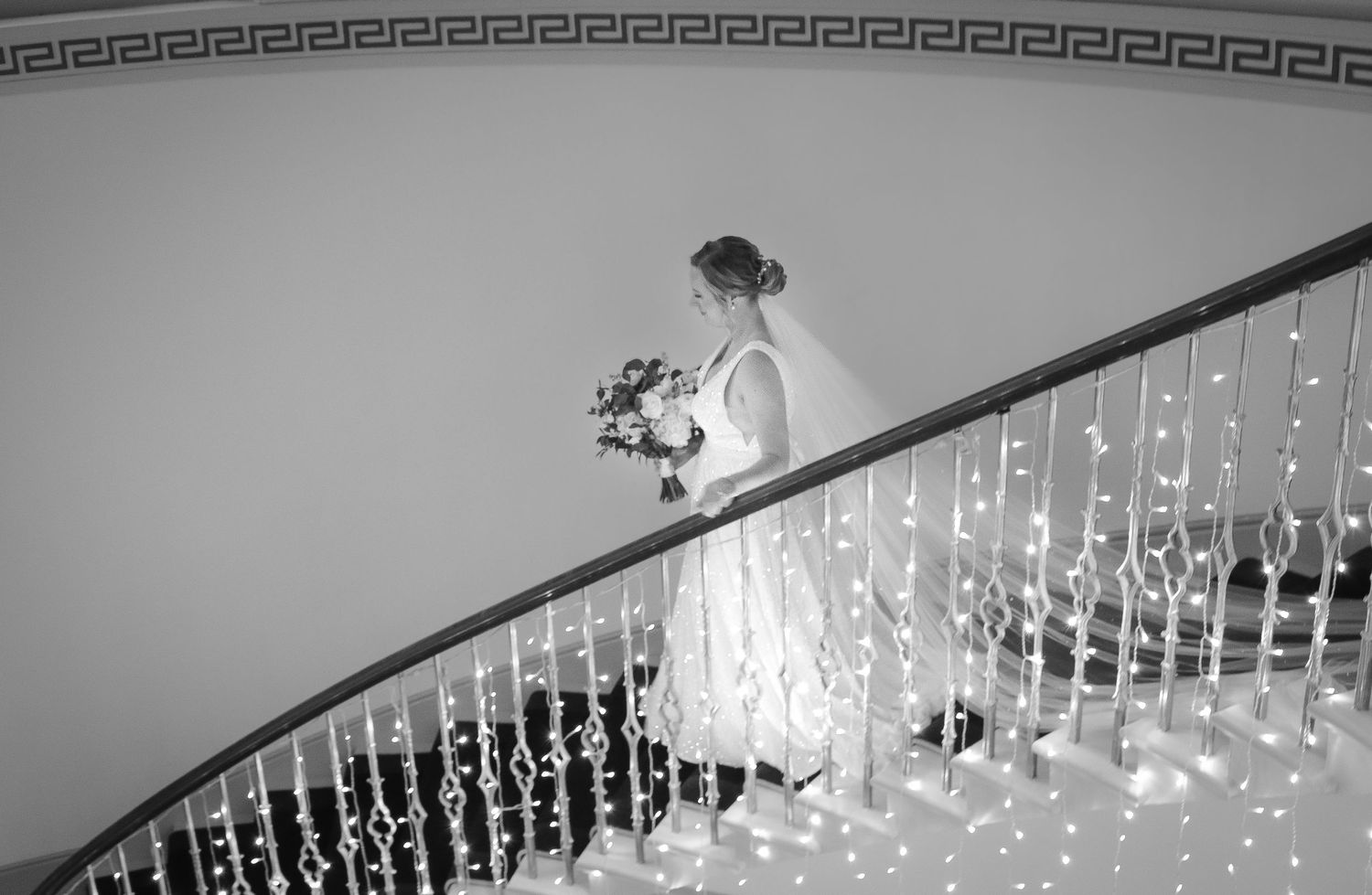 Elegant black and white image of someone in wedding attire descending a curved staircase adorned with twinkling lights.