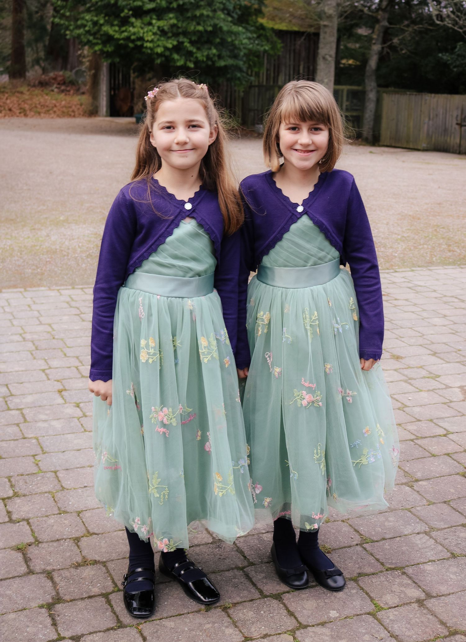 Two children in matching mint green dresses with floral designs and purple cardigans stand together on a brick pathway.