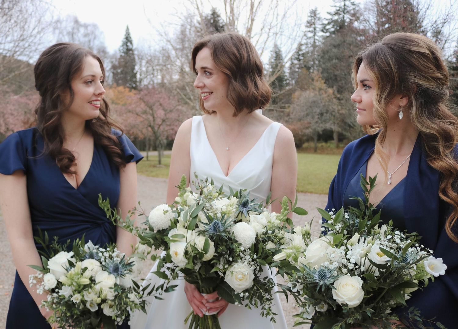 Three people hold elegant white and green floral bouquets while wearing navy and white dresses at an outdoor wedding.