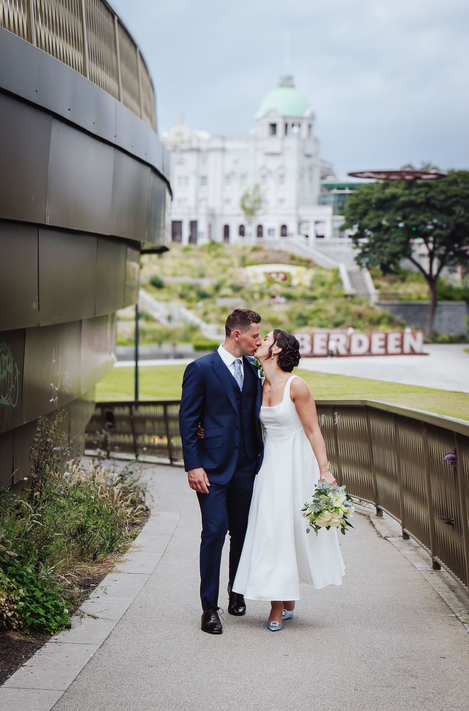 A couple in wedding attire walking together along a path with Aberdeen landmark building visible in background.