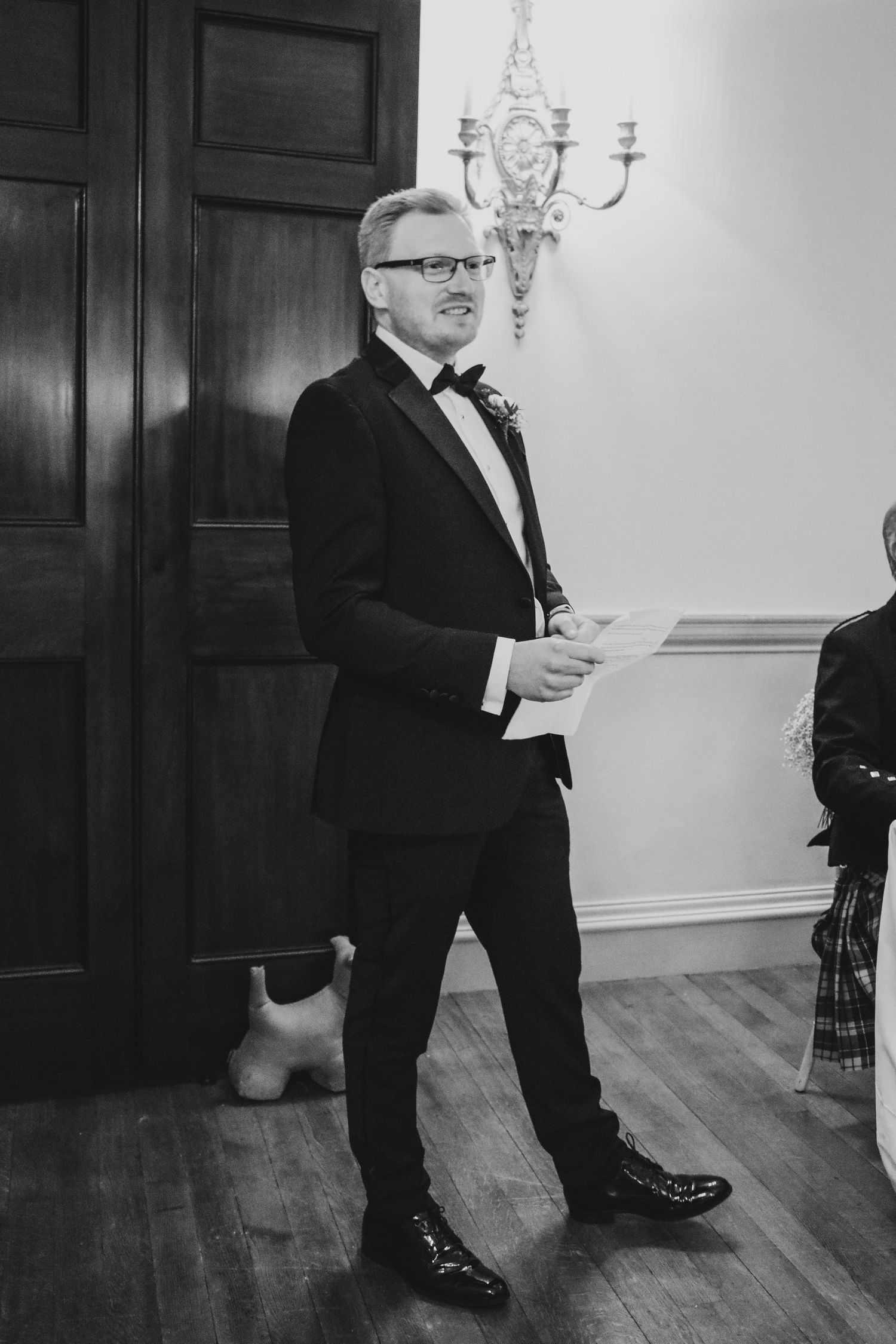Formal black and white portrait of person in tuxedo standing next to a decorative wall sconce at an elegant event.