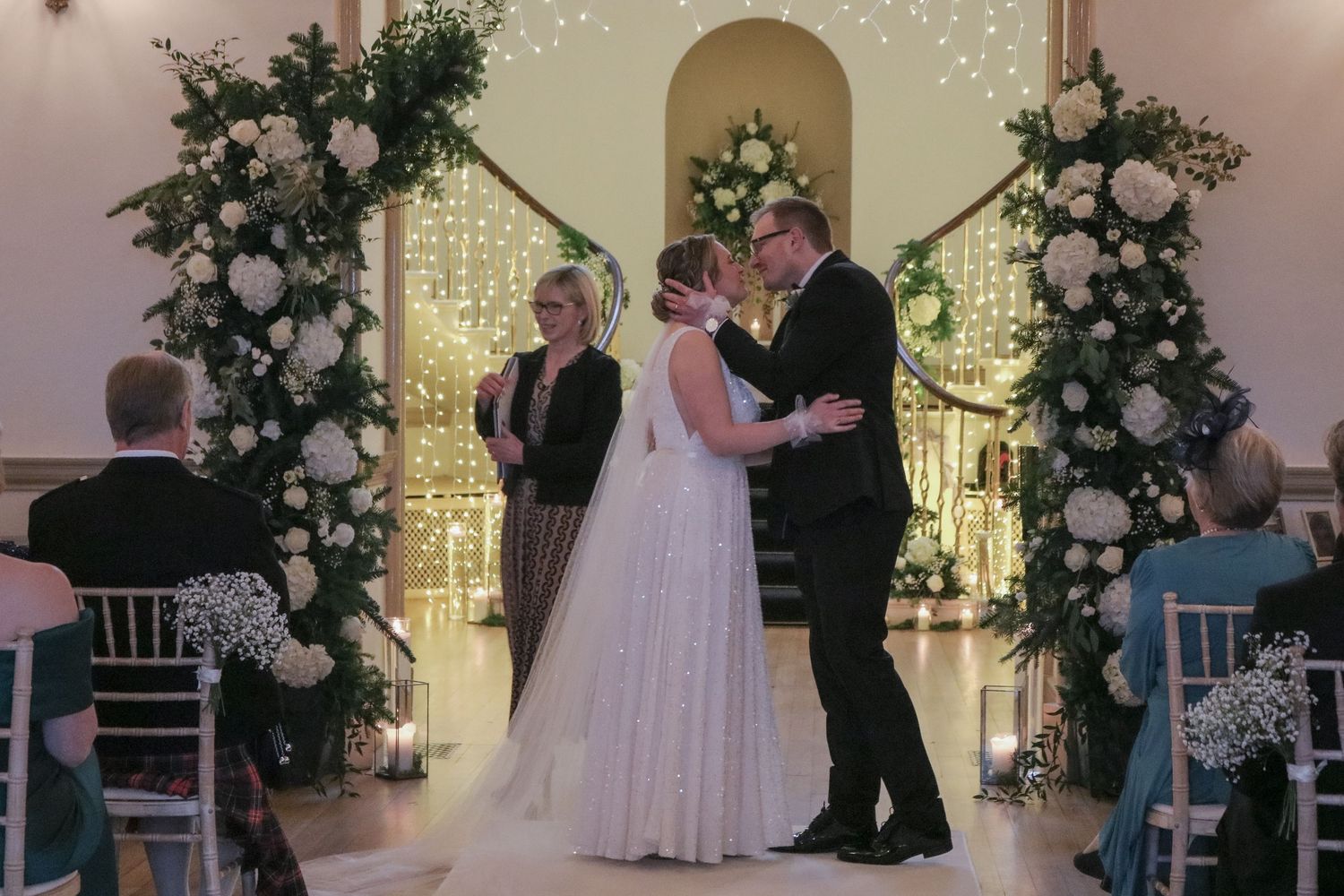 Indoor wedding ceremony featuring elegant white floral arches with glowing lights in a romantic ballroom setting.