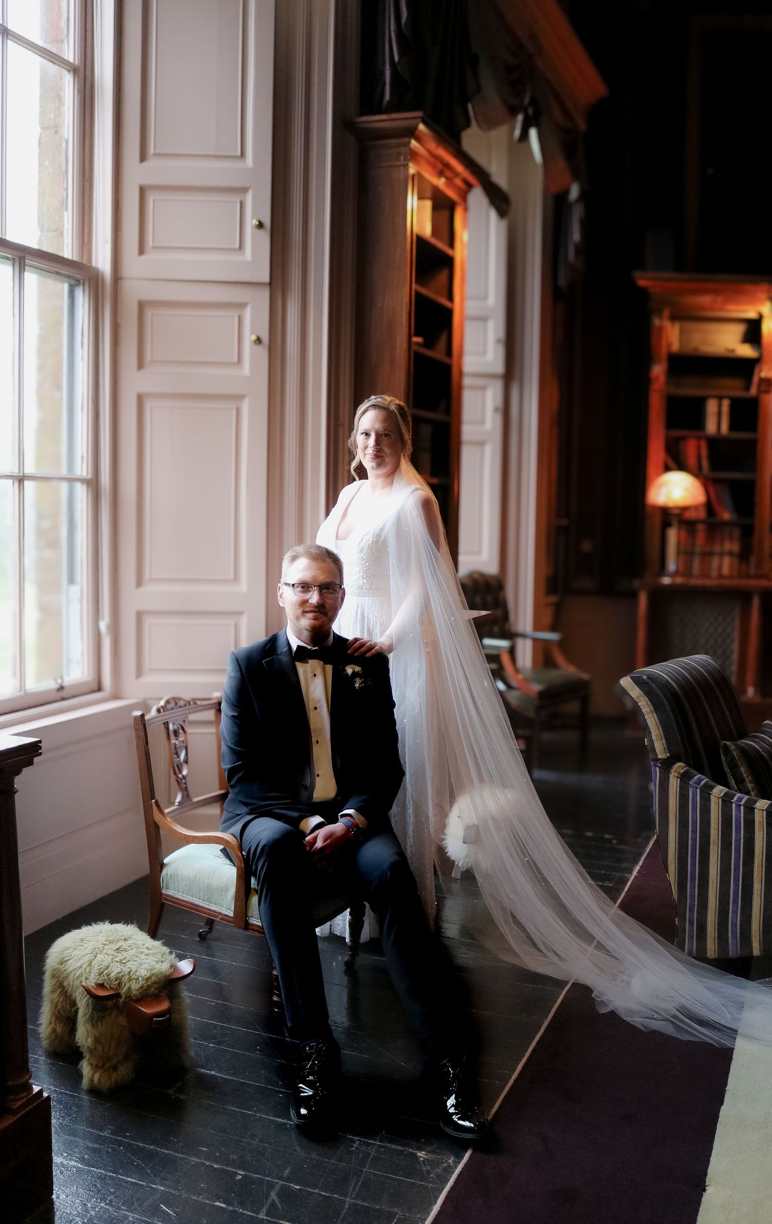A couple in wedding attire poses in an elegant room with tall windows and dark wood floors by vintage furniture.