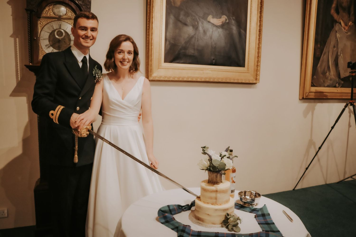 A happy couple in wedding attire stands together while cutting their rustic tiered wedding cake at an indoor reception.