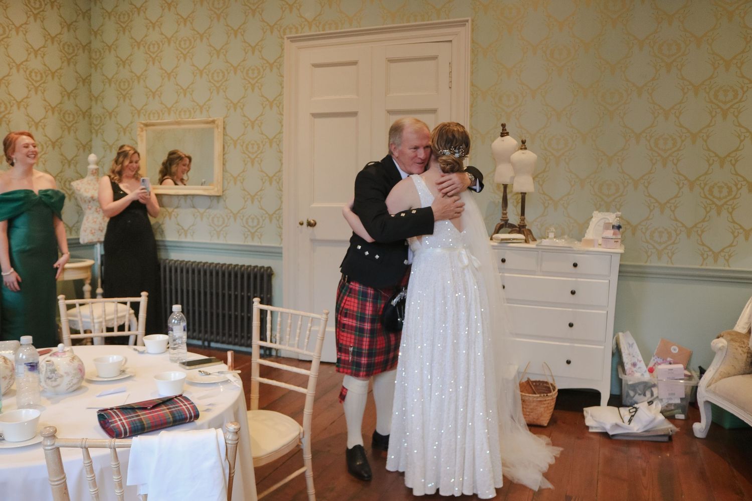 A couple embraces at an elegant wedding celebration in a vintage-styled room with yellow wallpaper and formal table settings.