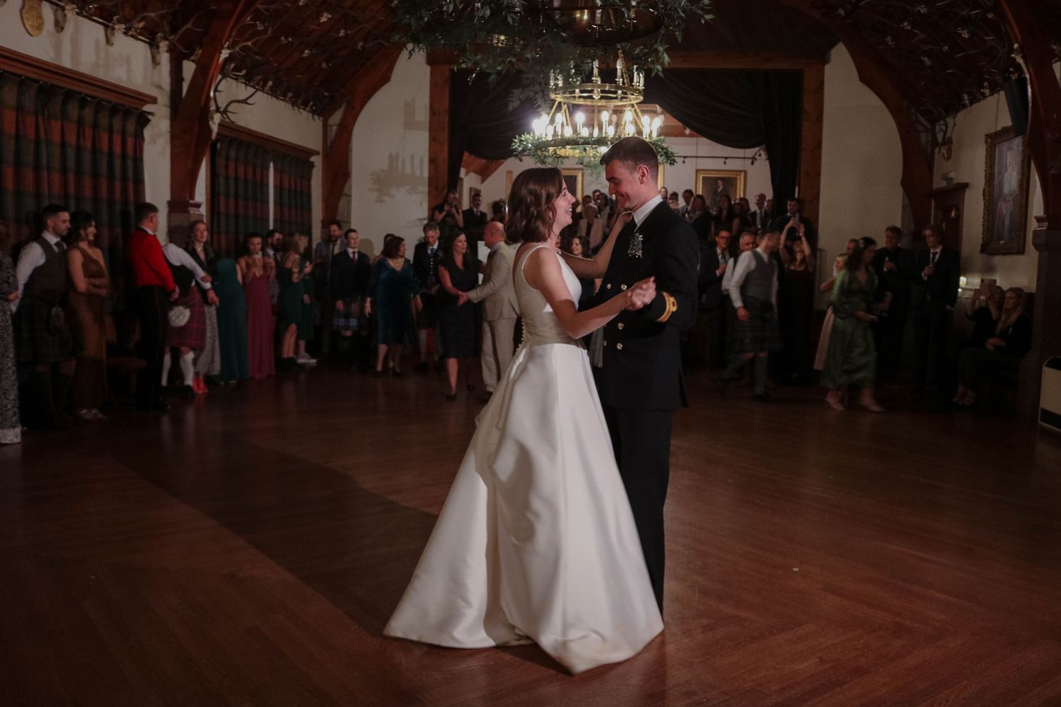 Two people share their first dance as newlyweds on a wooden dance floor in an elegant reception hall while guests watch.