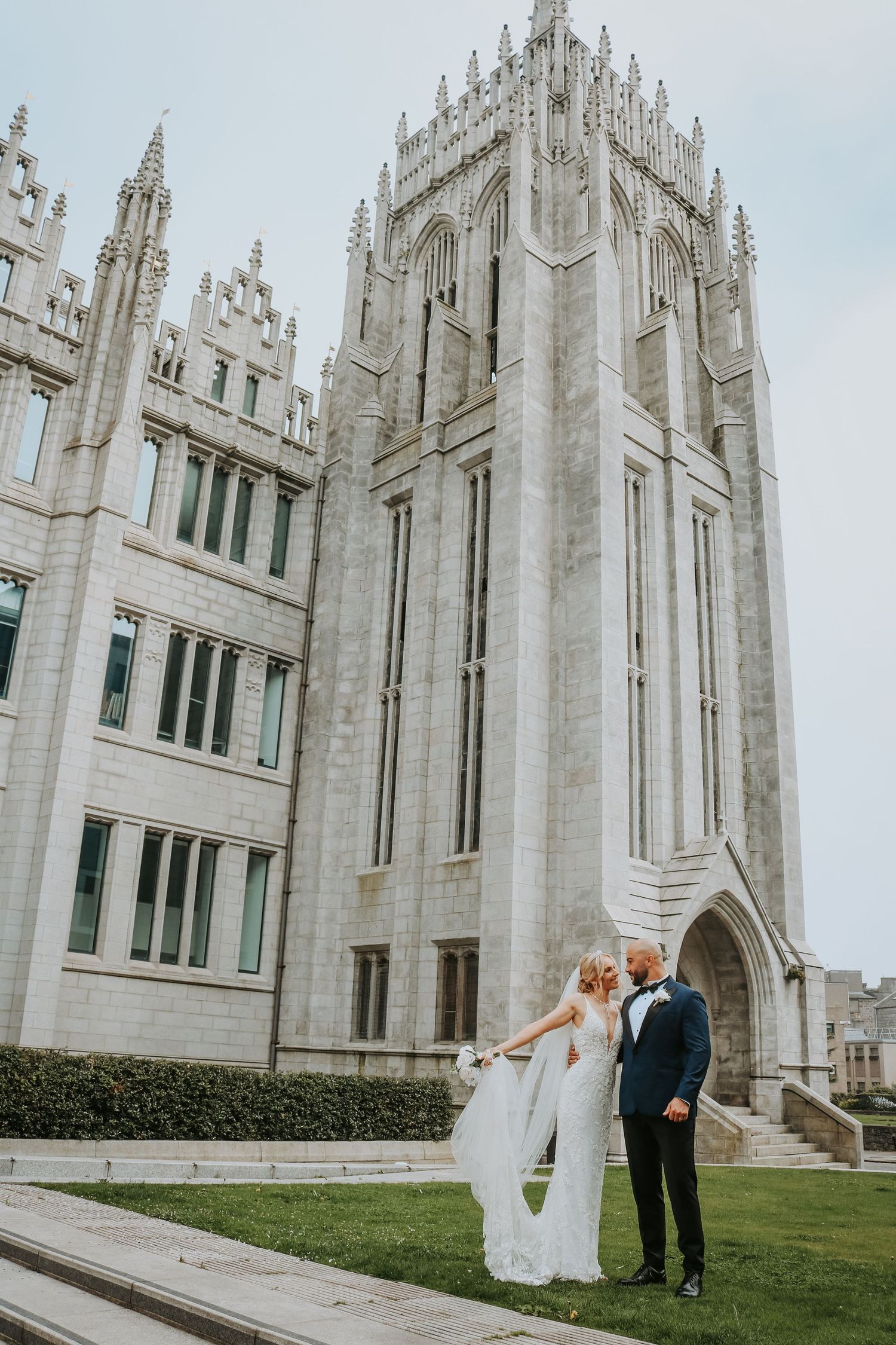 A wedding couple poses in front of a grand Gothic-style university building with white stone architecture.