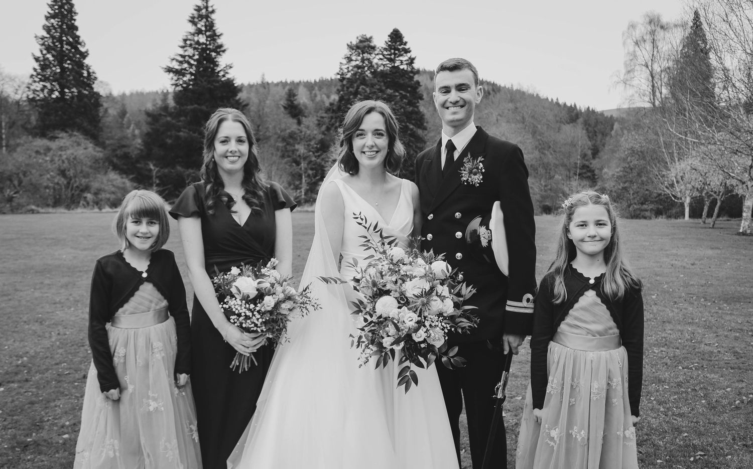 Black and white outdoor wedding photograph with a wedding party of five people on a hillside with evergreen trees.