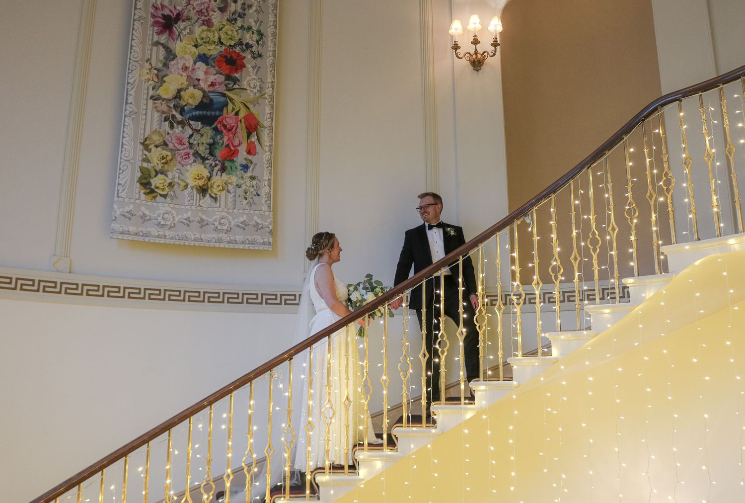 A couple descends an elegant white staircase adorned with twinkling lights and an ornate floral wall painting.