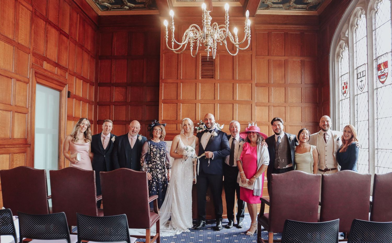 Wedding guests raise their glasses for a celebratory toast inside an elegant wood-paneled room with ornate windows and chandelier.