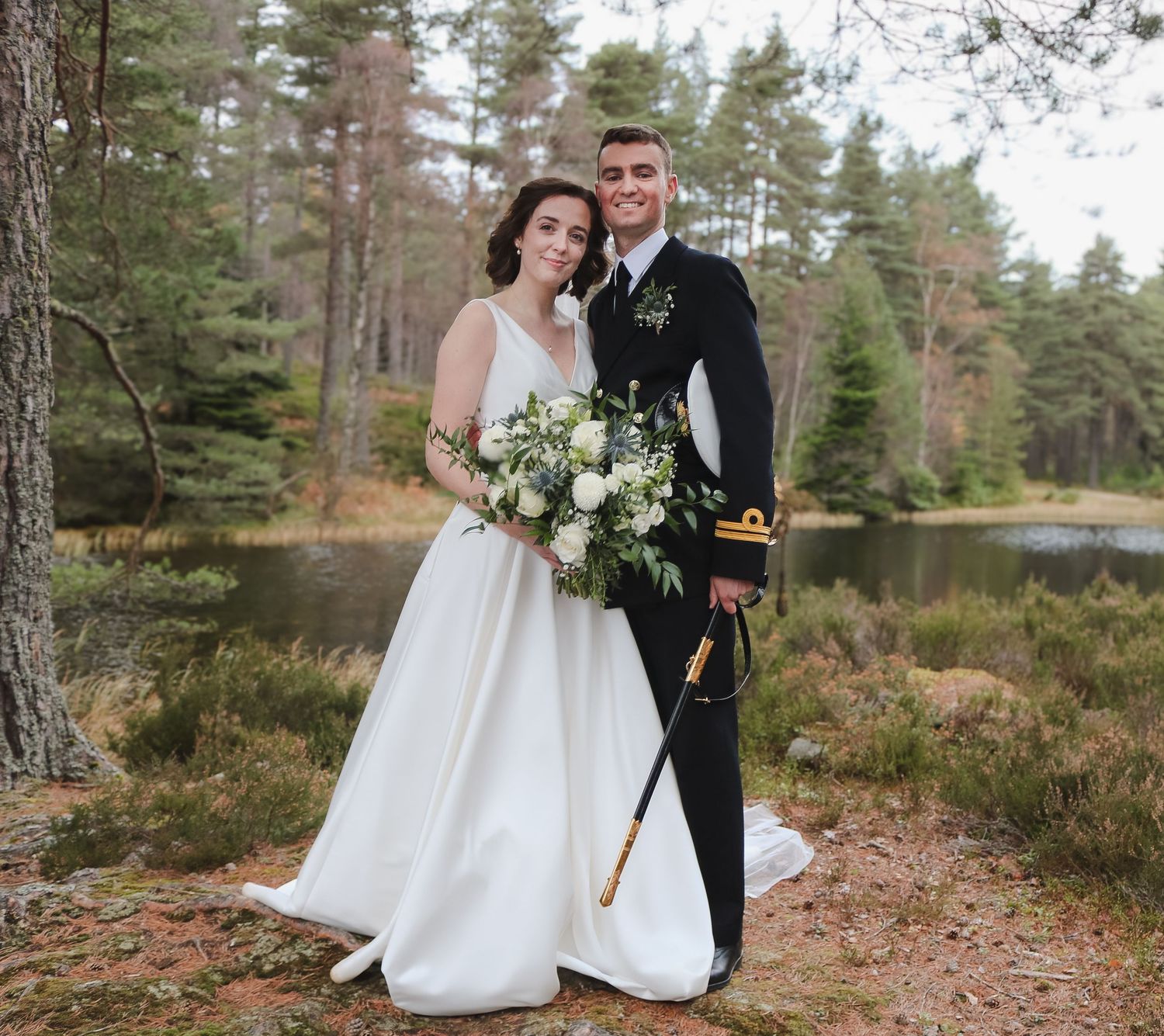 A wedding couple poses in military dress and white gown against evergreen forest and lake backdrop on an autumn day.