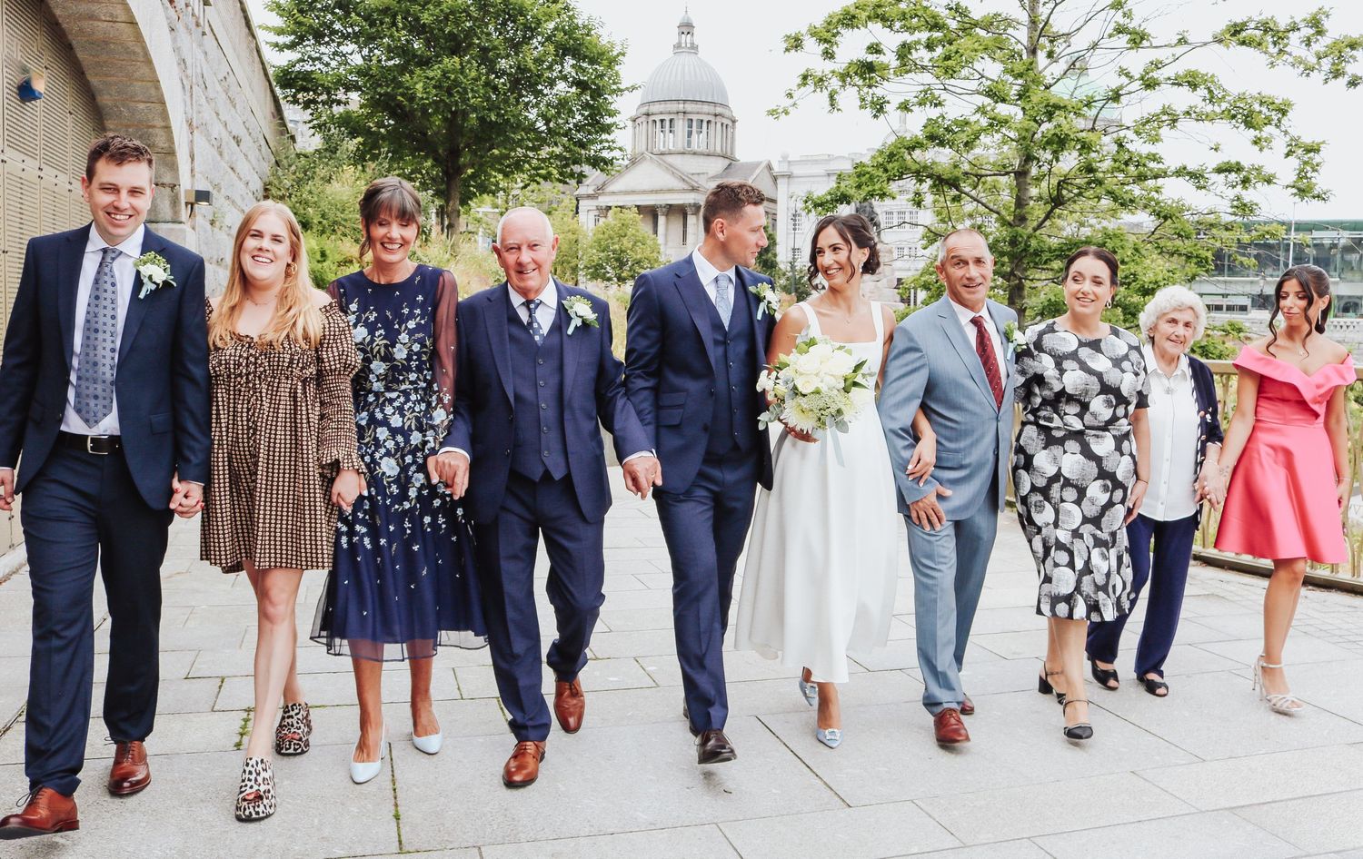 Wedding party in formal attire poses together on sidewalk with domed building visible behind them.