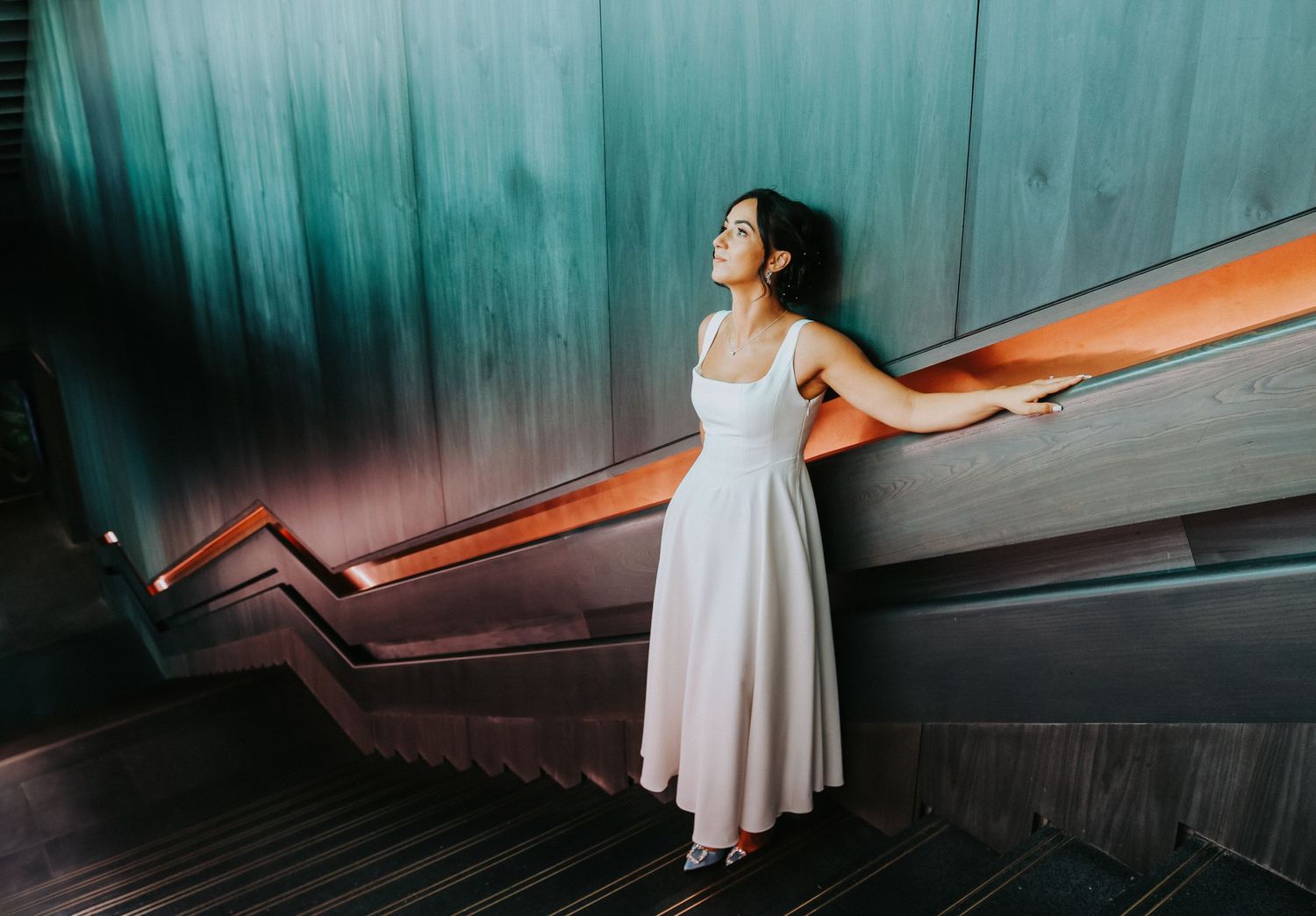 Person in a flowy white dress poses gracefully against teal walls on a modern staircase with orange railings.