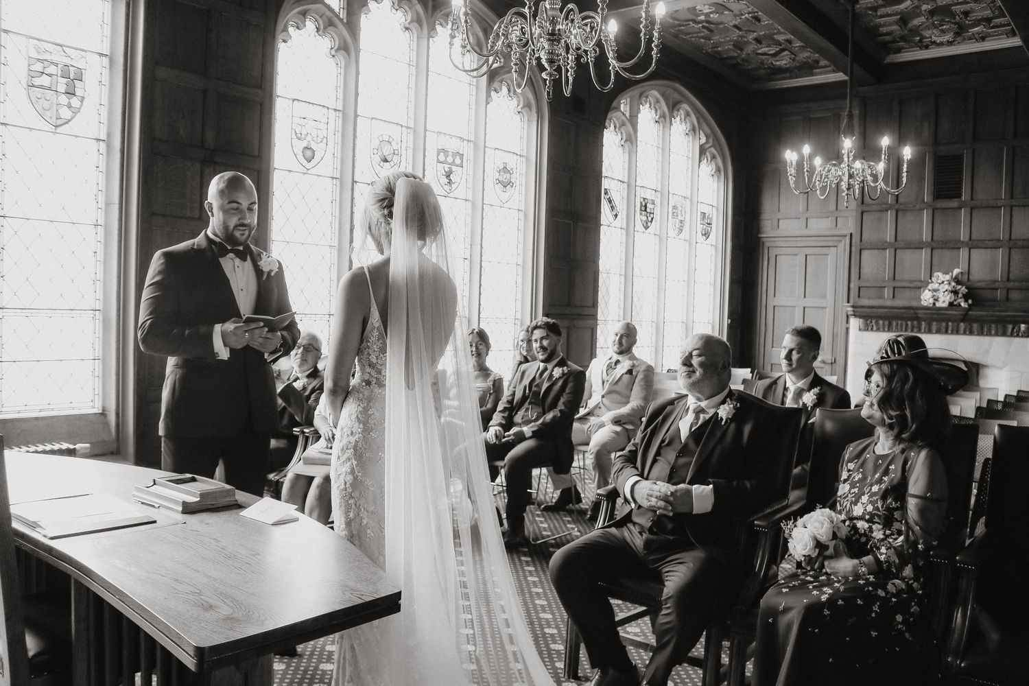 Black and white photo of a wedding ceremony in a grand hall with tall windows and ornate details.