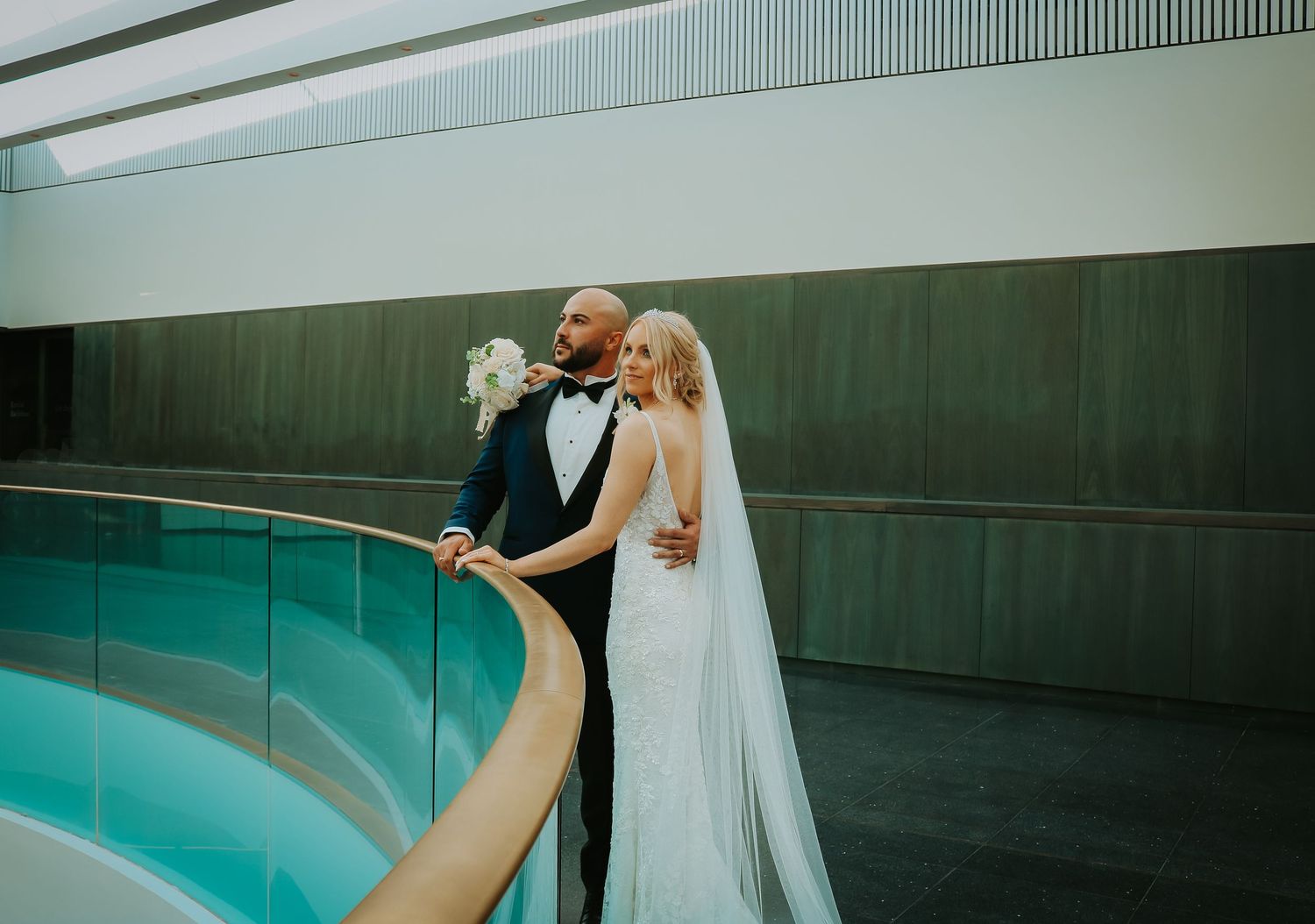 A couple poses elegantly beside a turquoise indoor pool, with the bride wearing a flowing white veil.