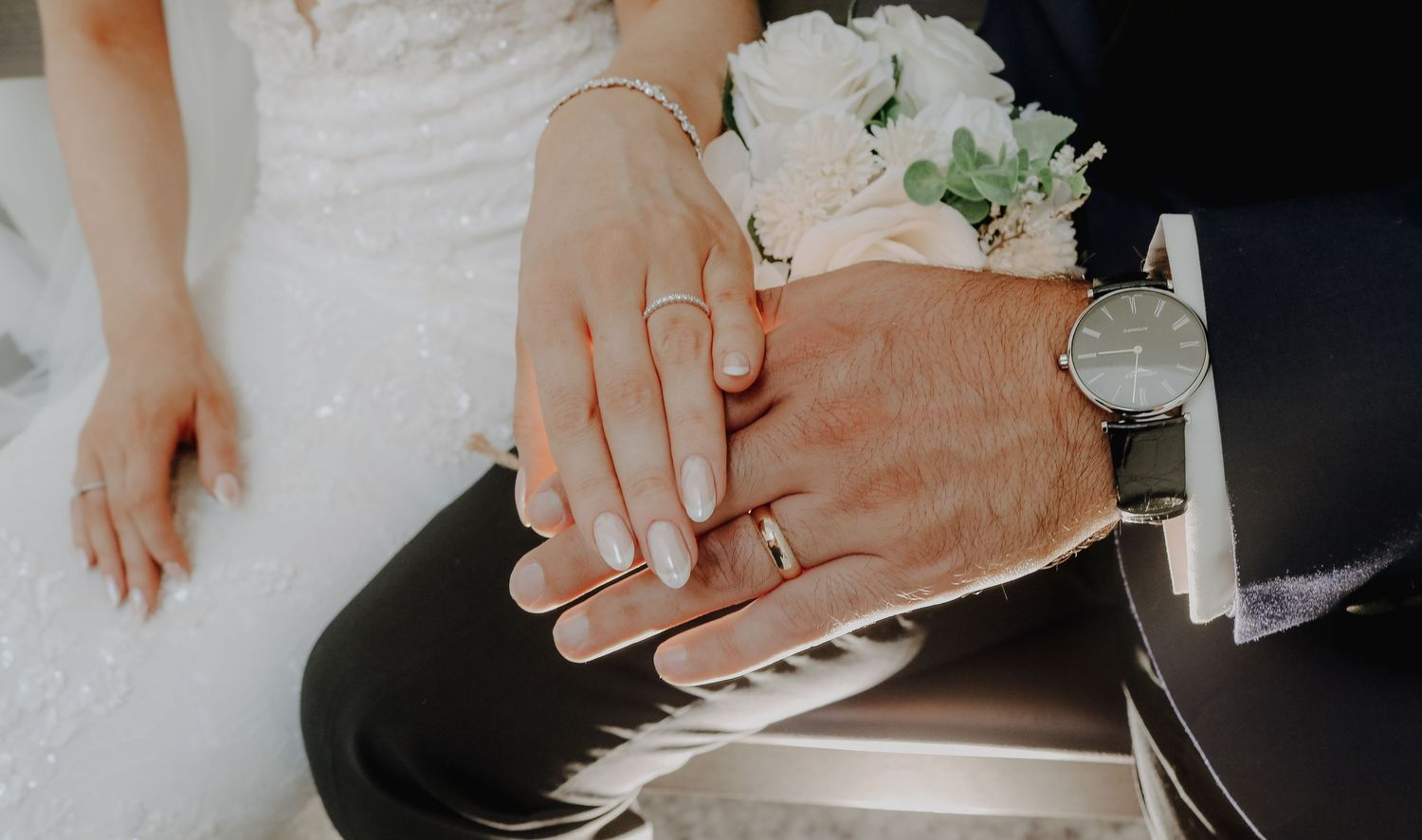 Close-up of bride and groom's hands with wedding rings resting on white flowers.