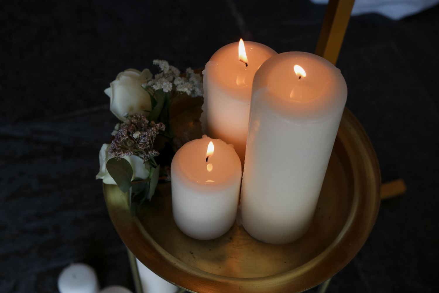 Three lit white pillar candles glowing on a gold plate with small white flowers against a dark background.