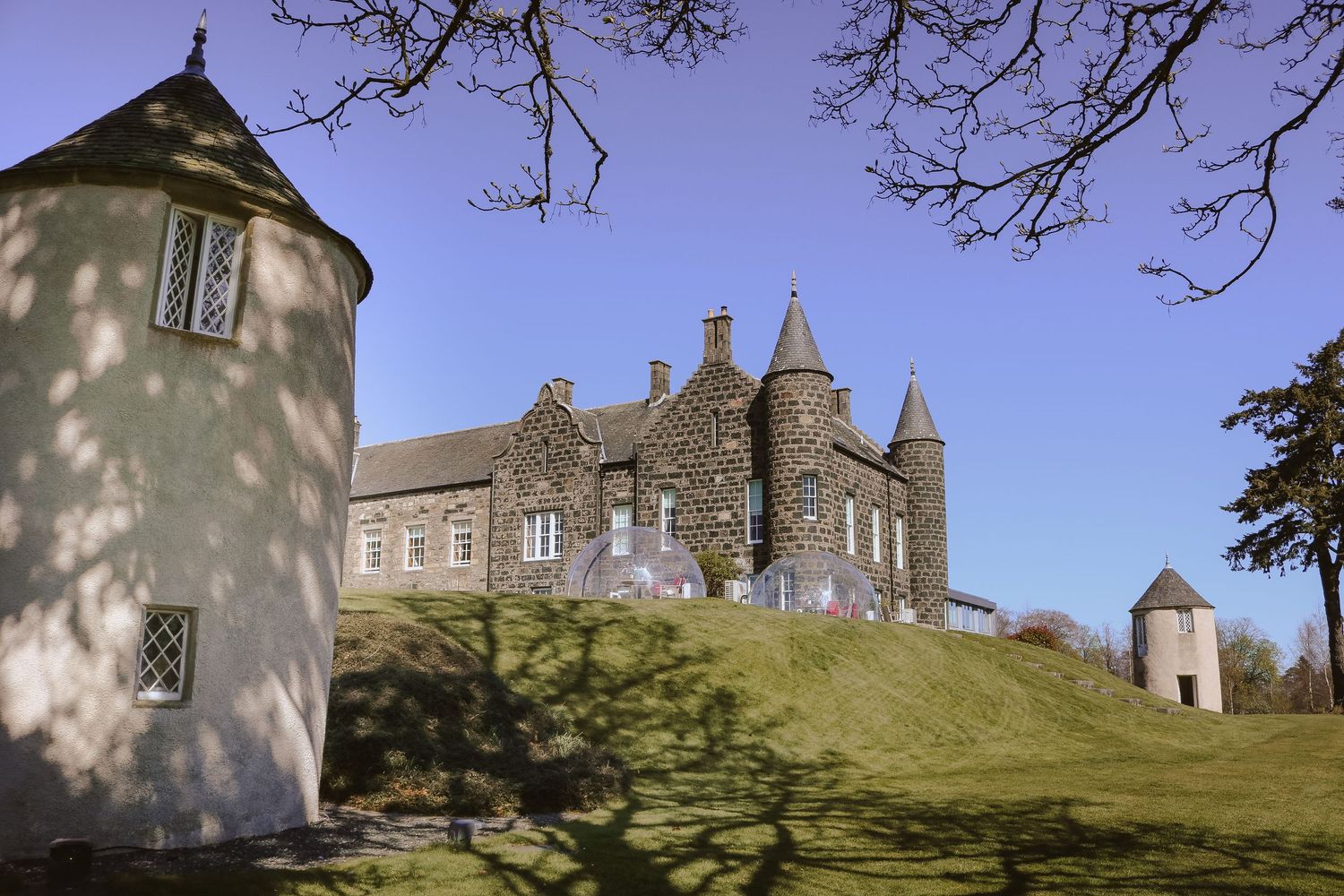 Historic stone castle with turrets sits atop grassy hill with white towers in foreground against blue sky.
