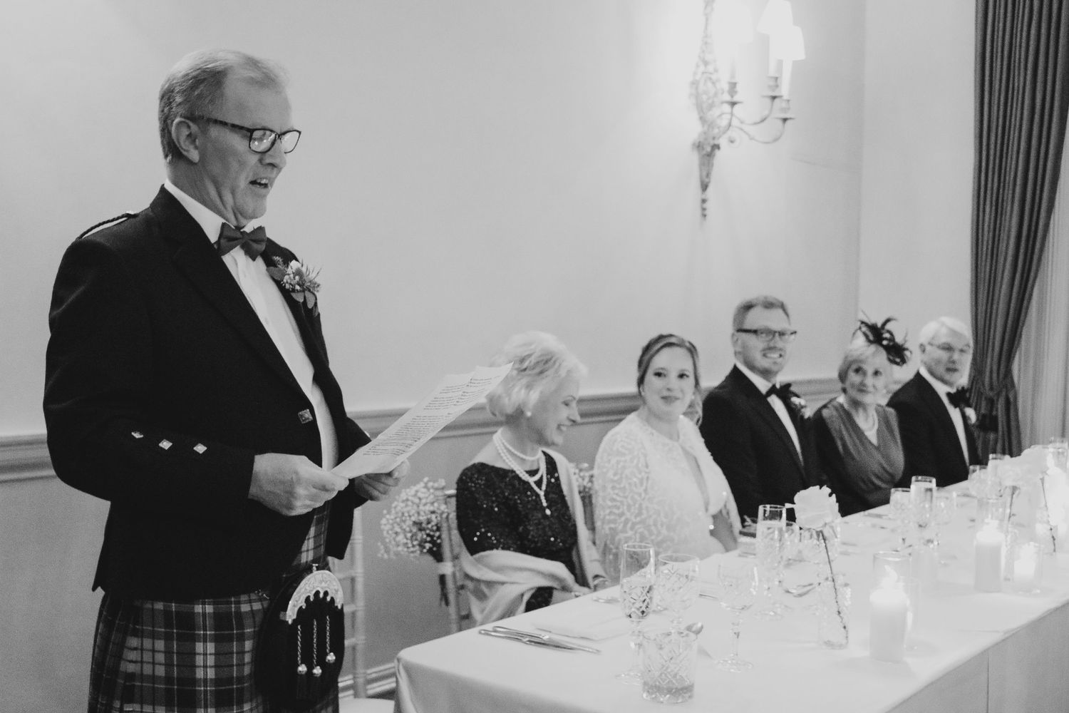 Black and white photo of a wedding reception toast at head table with speaker wearing traditional Scottish kilt.