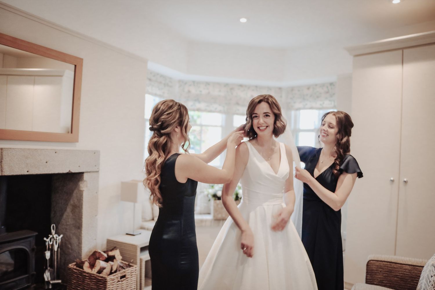 Bridesmaids in black dresses help adjust a white wedding gown in a bright room with a fireplace.