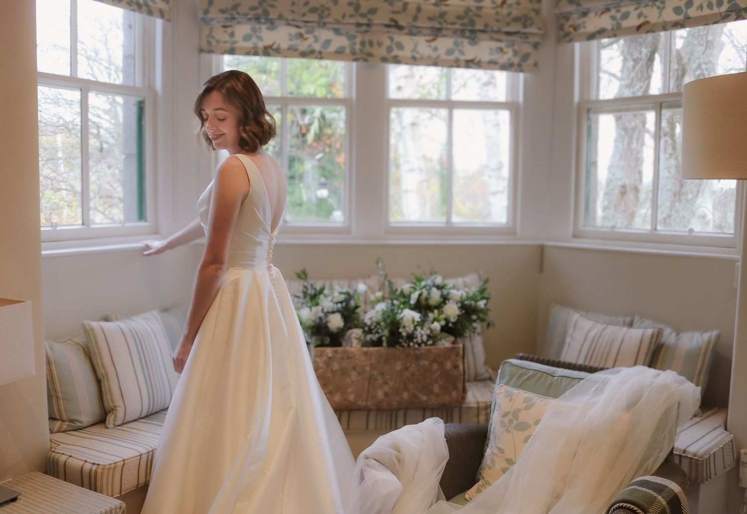 A flowing white wedding dress catches the light in a sunny window-filled room with plush seating and floral arrangements.