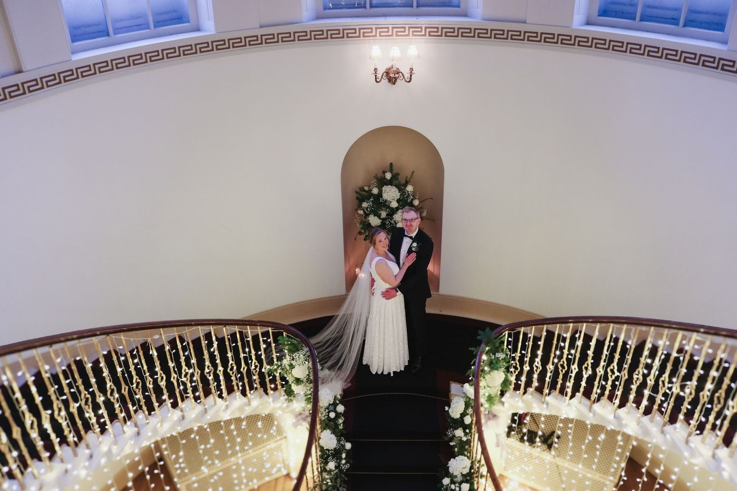 A couple stands together at the top of an elegant curved staircase decorated with flowers and warm lighting.