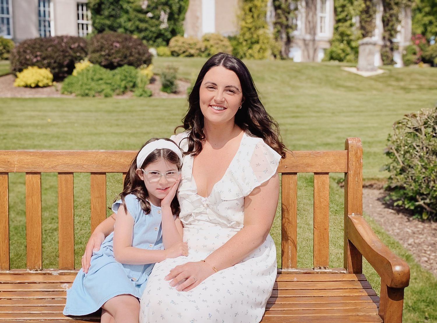 A parent and child sitting together on a wooden park bench in a lush garden setting on a sunny day.