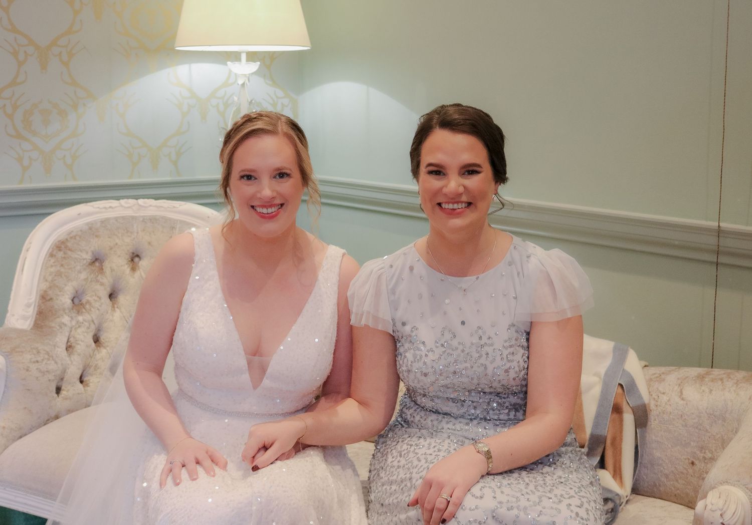 Two women in elegant dresses sitting together on a cream-colored tufted sofa in an elegant room setting.