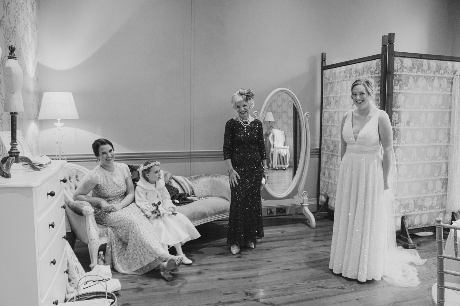 A vintage black and white photograph showing people in formal attire gathered in an elegant bedroom with period furniture.
