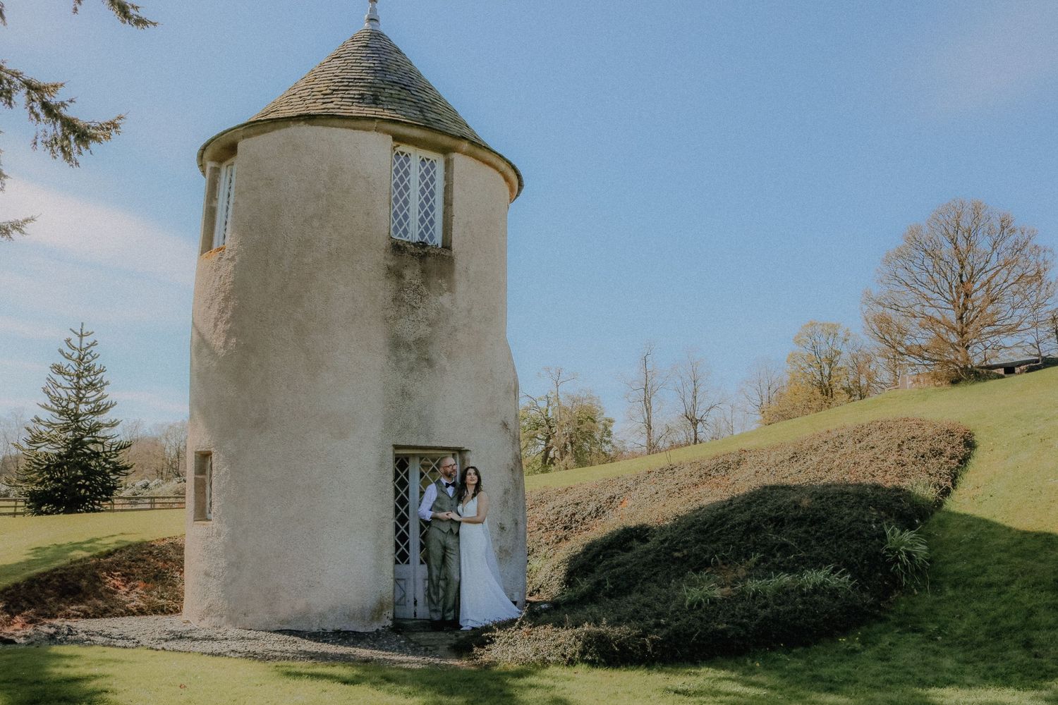 A white cylindrical tower with a conical roof stands on a green hillside against a blue sky on a sunny day.