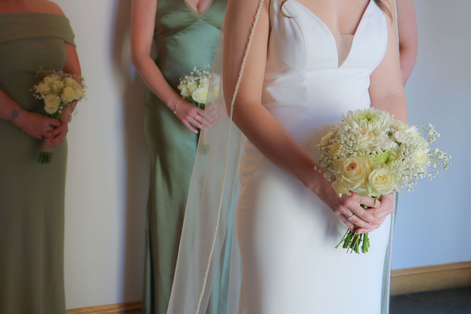 Bridesmaids in sage green dresses and bride in white hold delicate bouquets of white and green flowers.