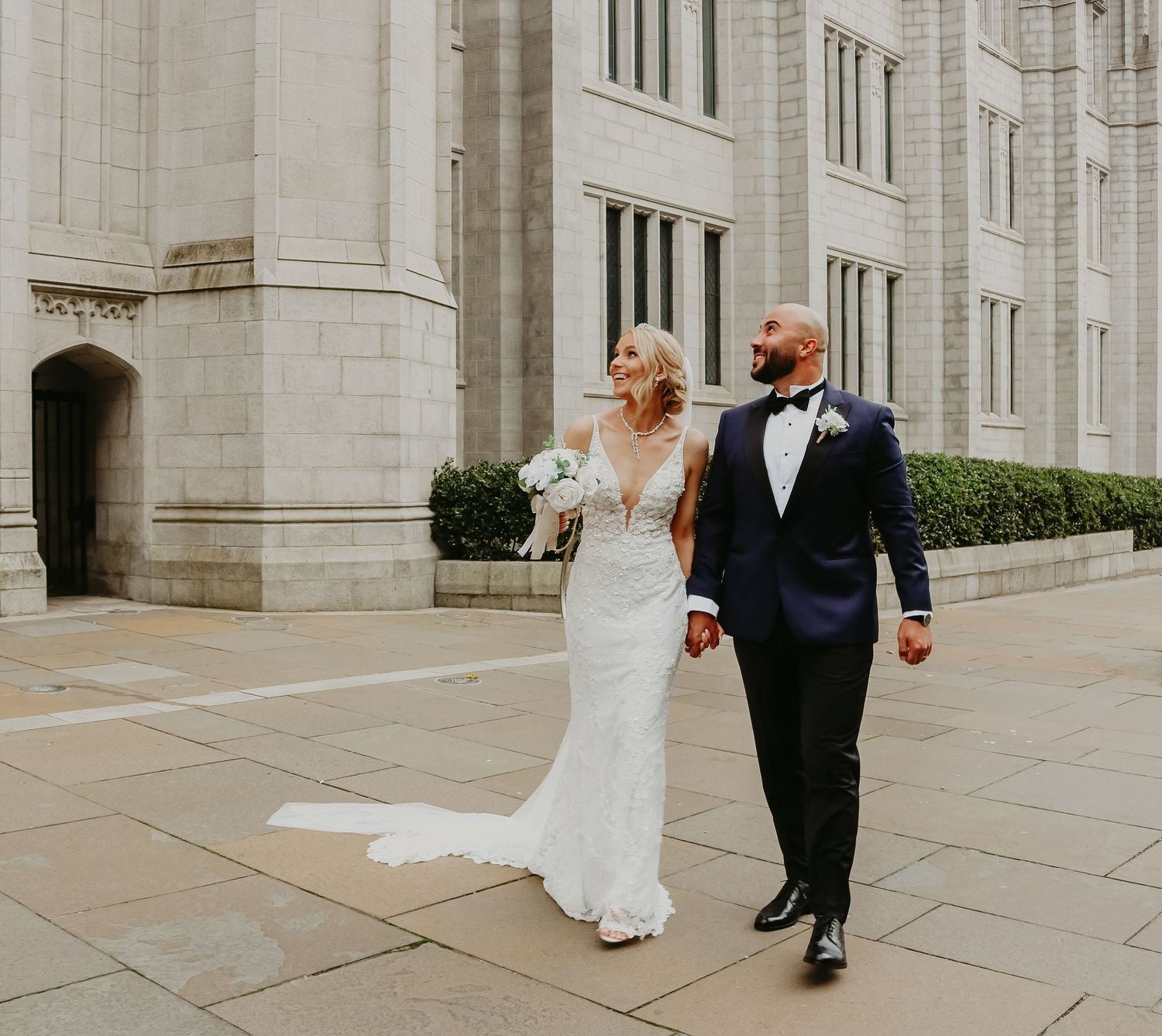 Newlyweds walk together outside a grand stone building with architectural details.