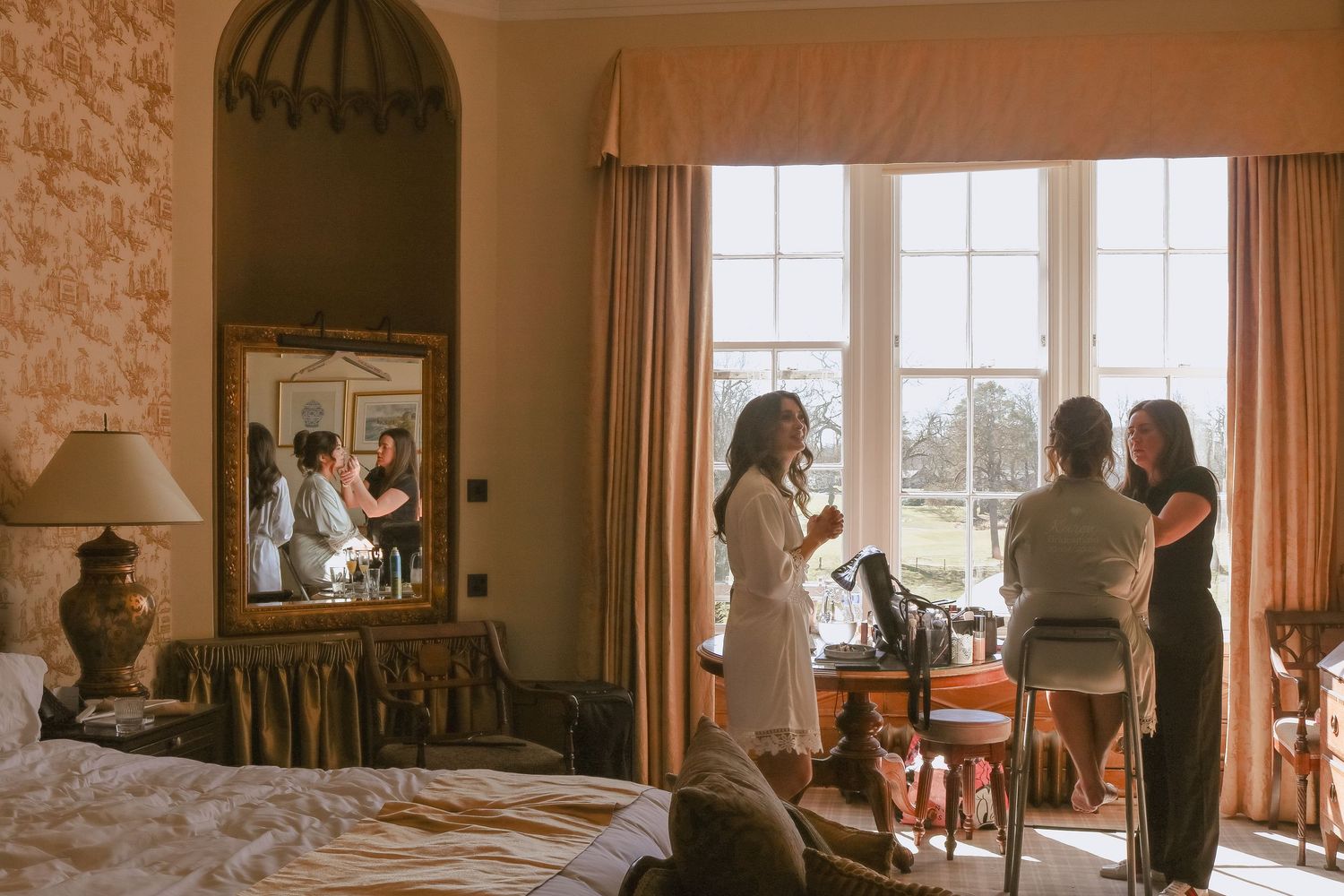 Light-filled bedroom interior with natural decor showing gathering scene by windows and reflection in vanity mirror.