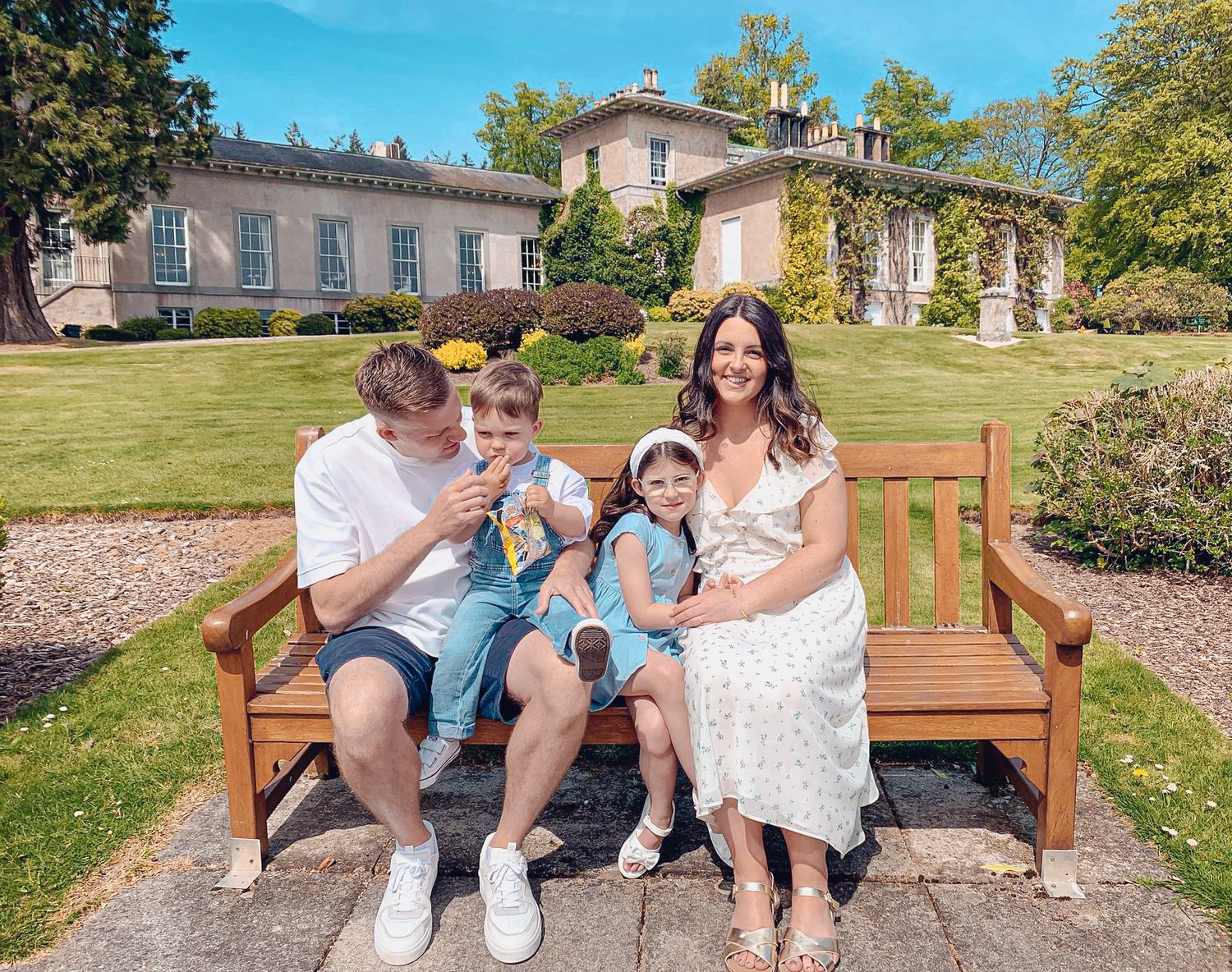 Family relaxing together on wooden garden bench against backdrop of stately manor house with ivy-covered walls.