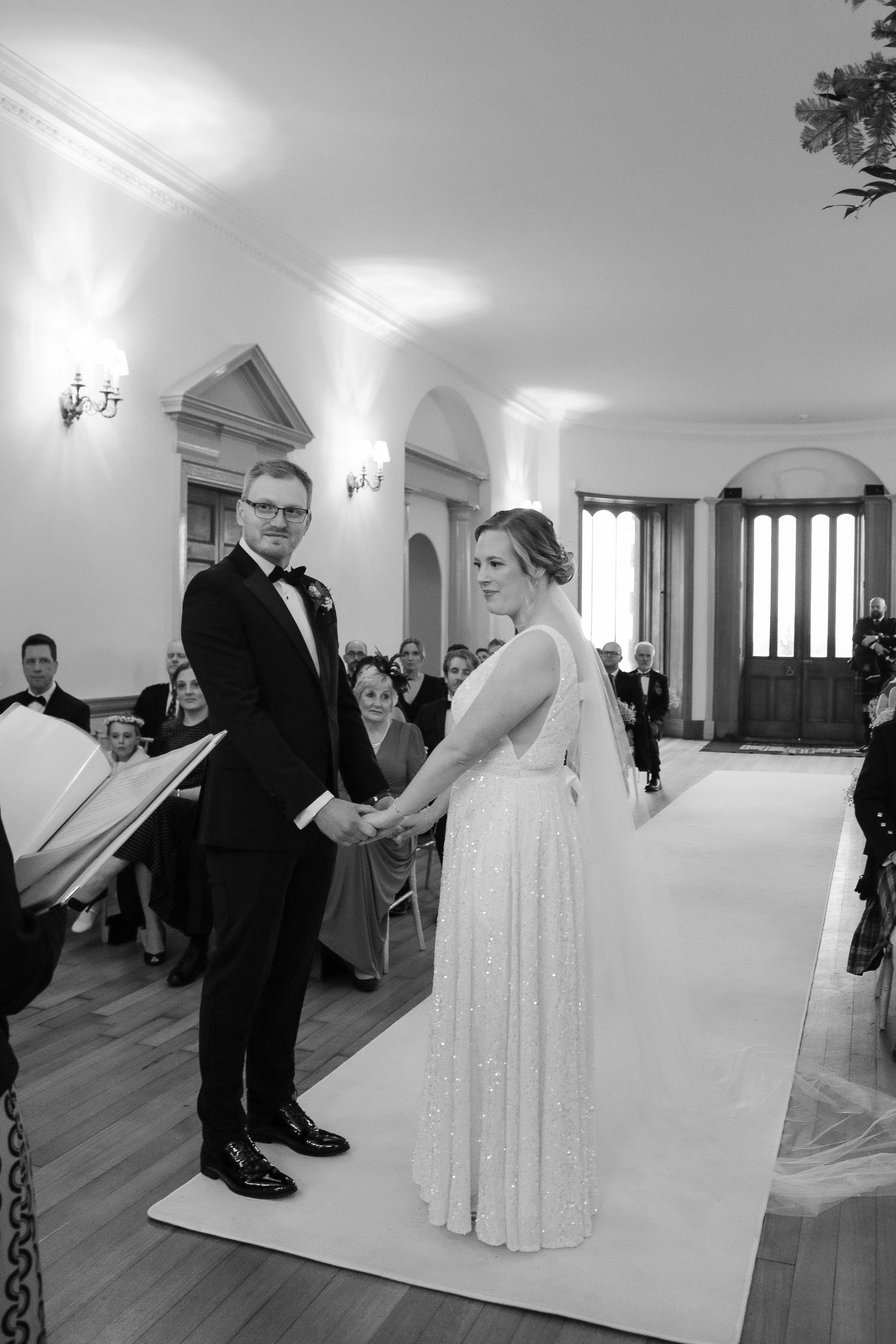 A wedding ceremony taking place in an elegant white room with arched doorways and wall sconces.