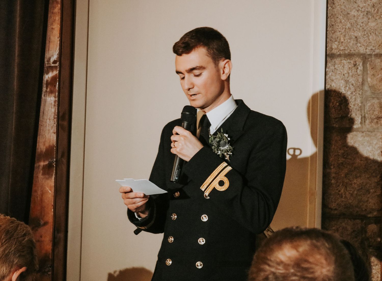 Someone in a dark formal coat delivers a wedding speech while holding a microphone and notes at an elegant venue.