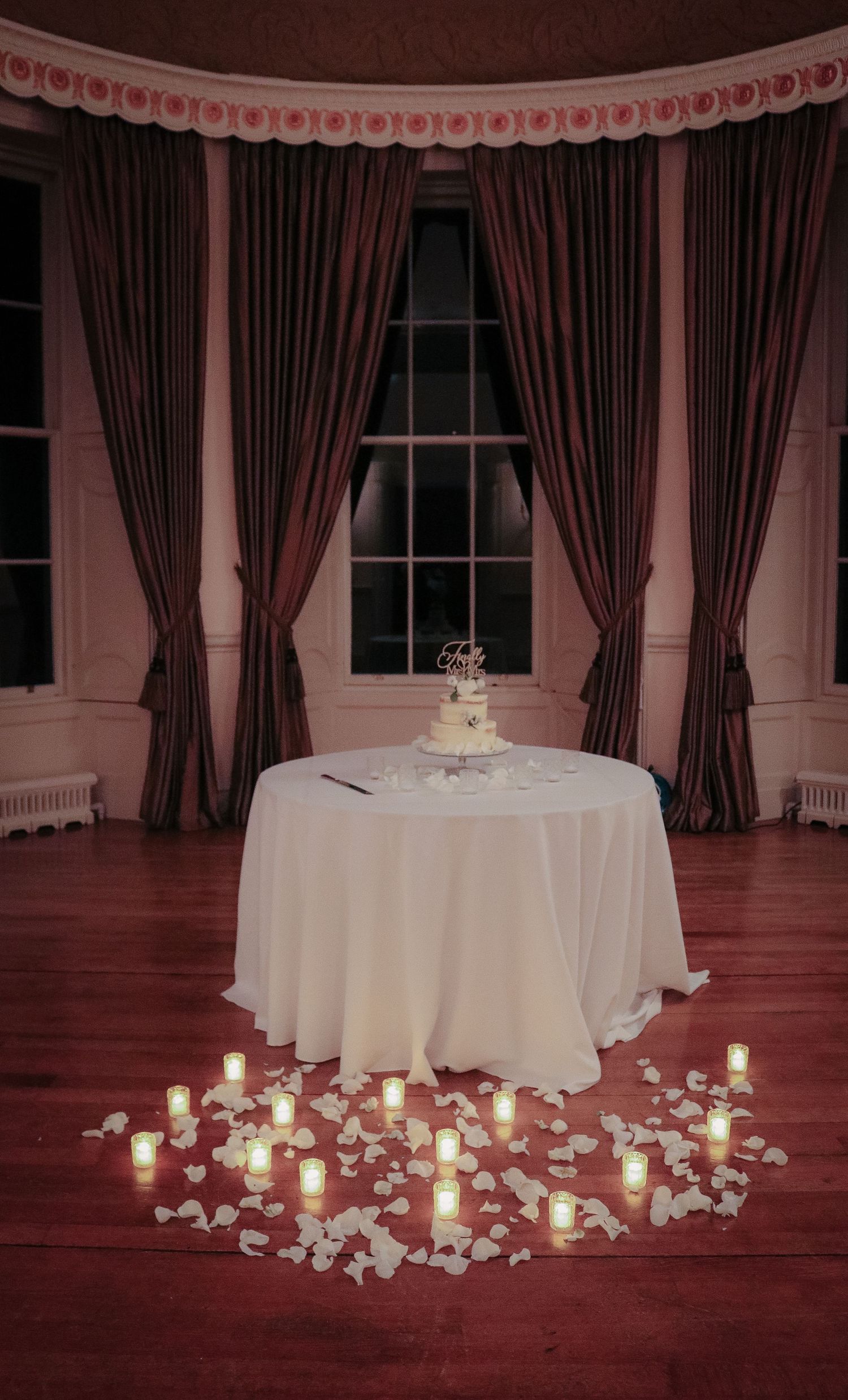A wedding cake sits on a white tablecloth surrounded by glowing candles and rose petals in an elegant bay window setting.