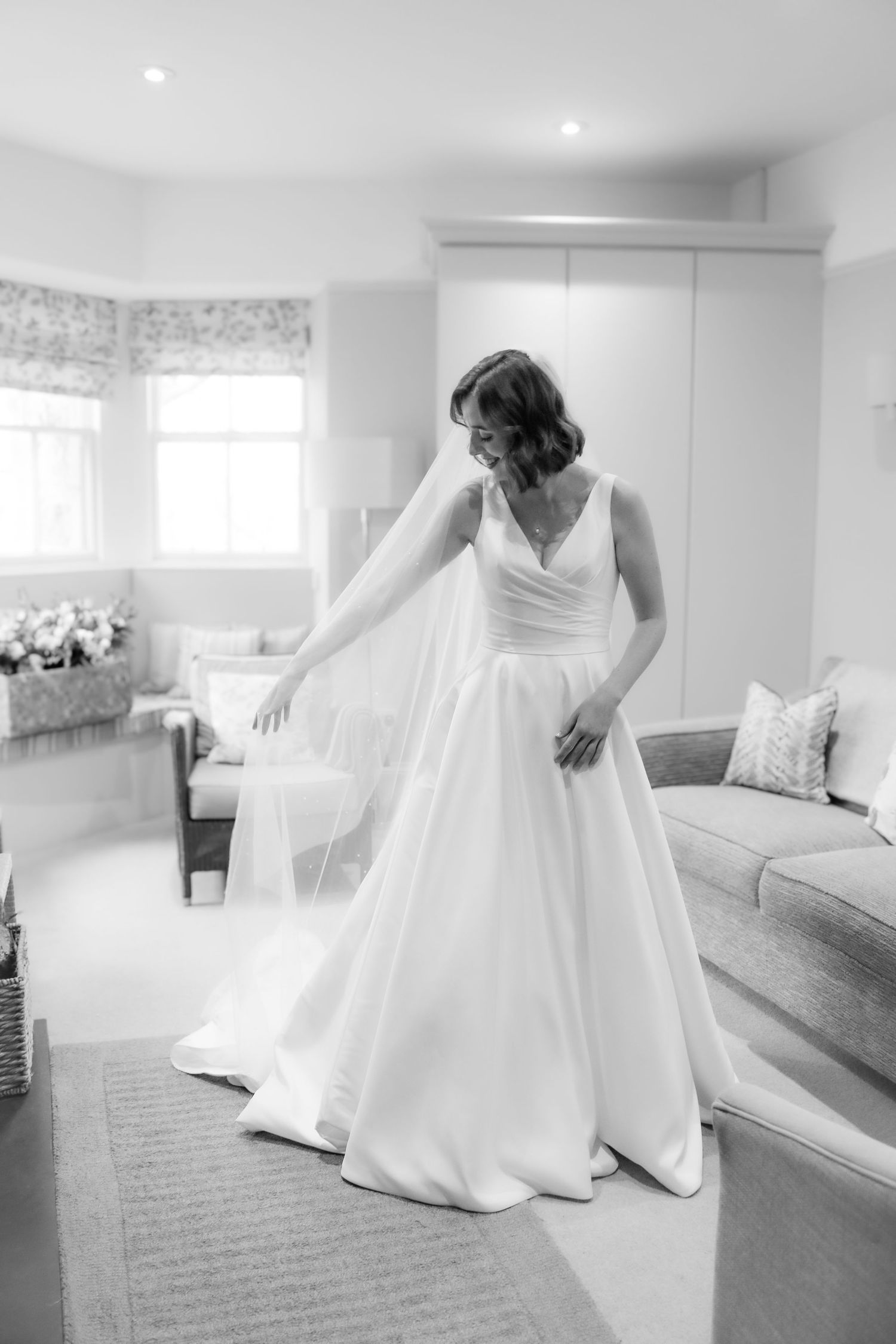 Black and white photo of a bride in an elegant V-neck wedding gown with flowy skirt in a bright room with windows.