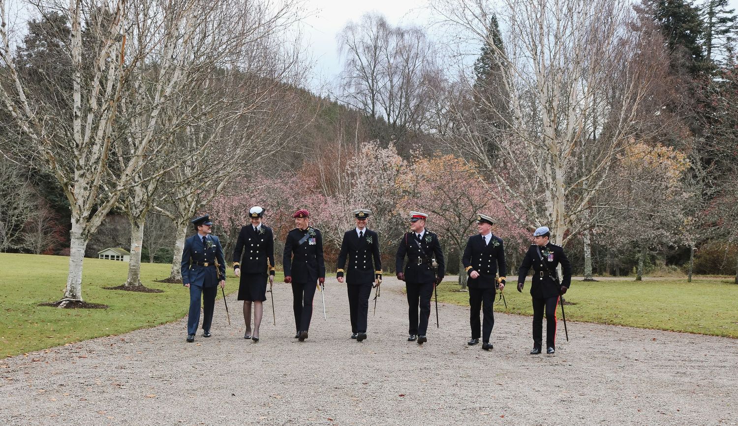 A group of uniformed military personnel walk together on a gravel path lined with bare trees in winter.