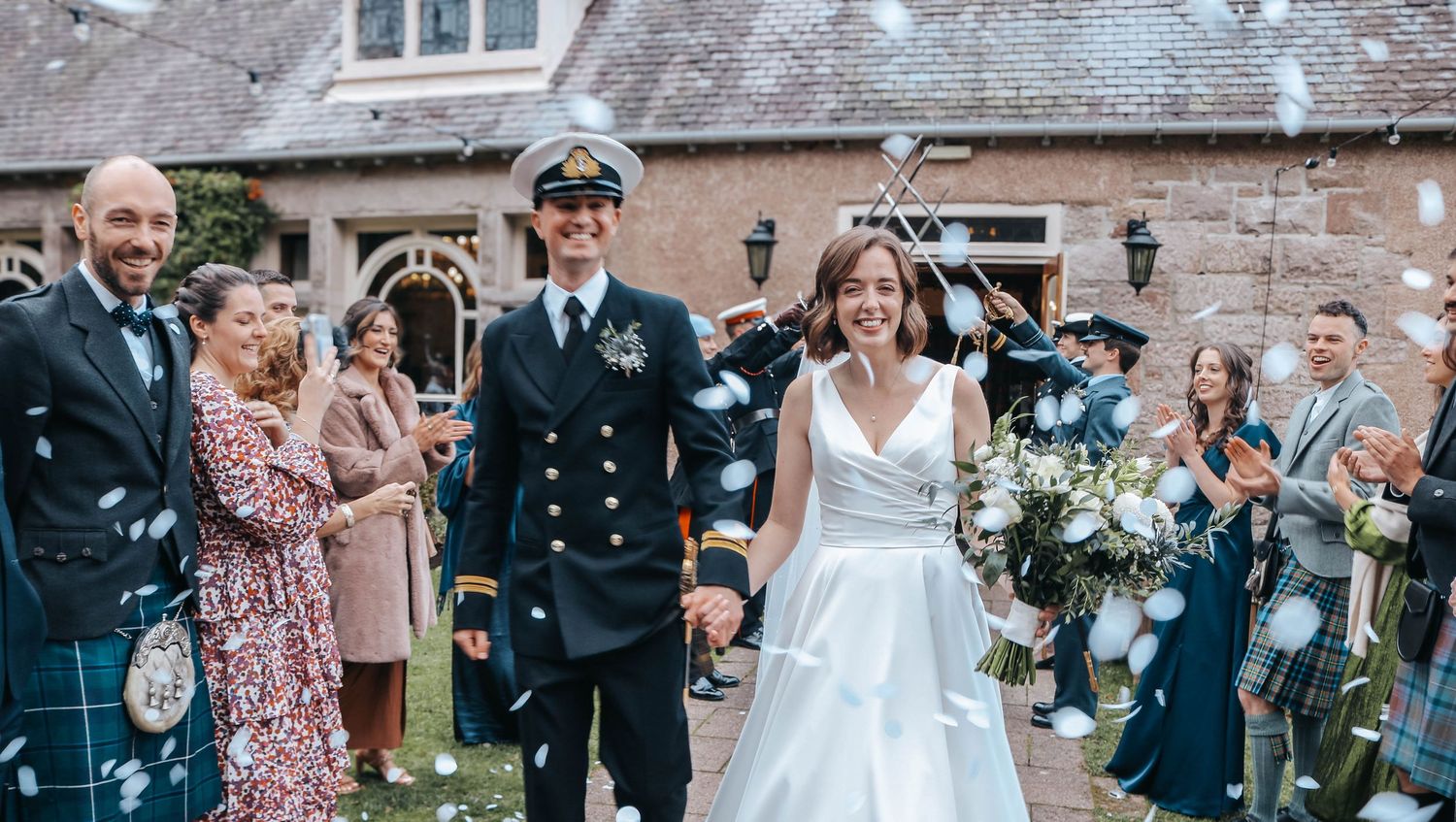 Wedding celebration outside stone building with people in formal attire, kilts, and military dress uniform throwing confetti.