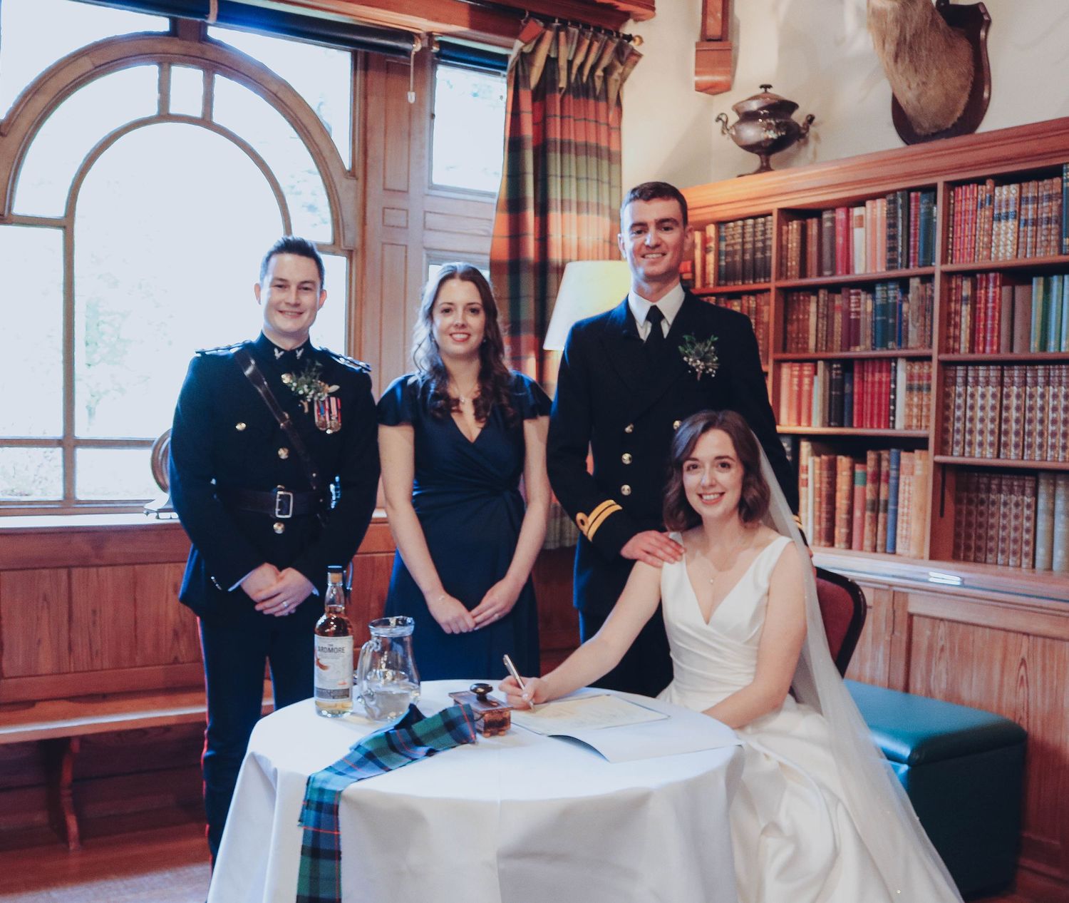 A formal group stands in a library room with wooden bookshelves and an arched window as someone signs documents at a table.