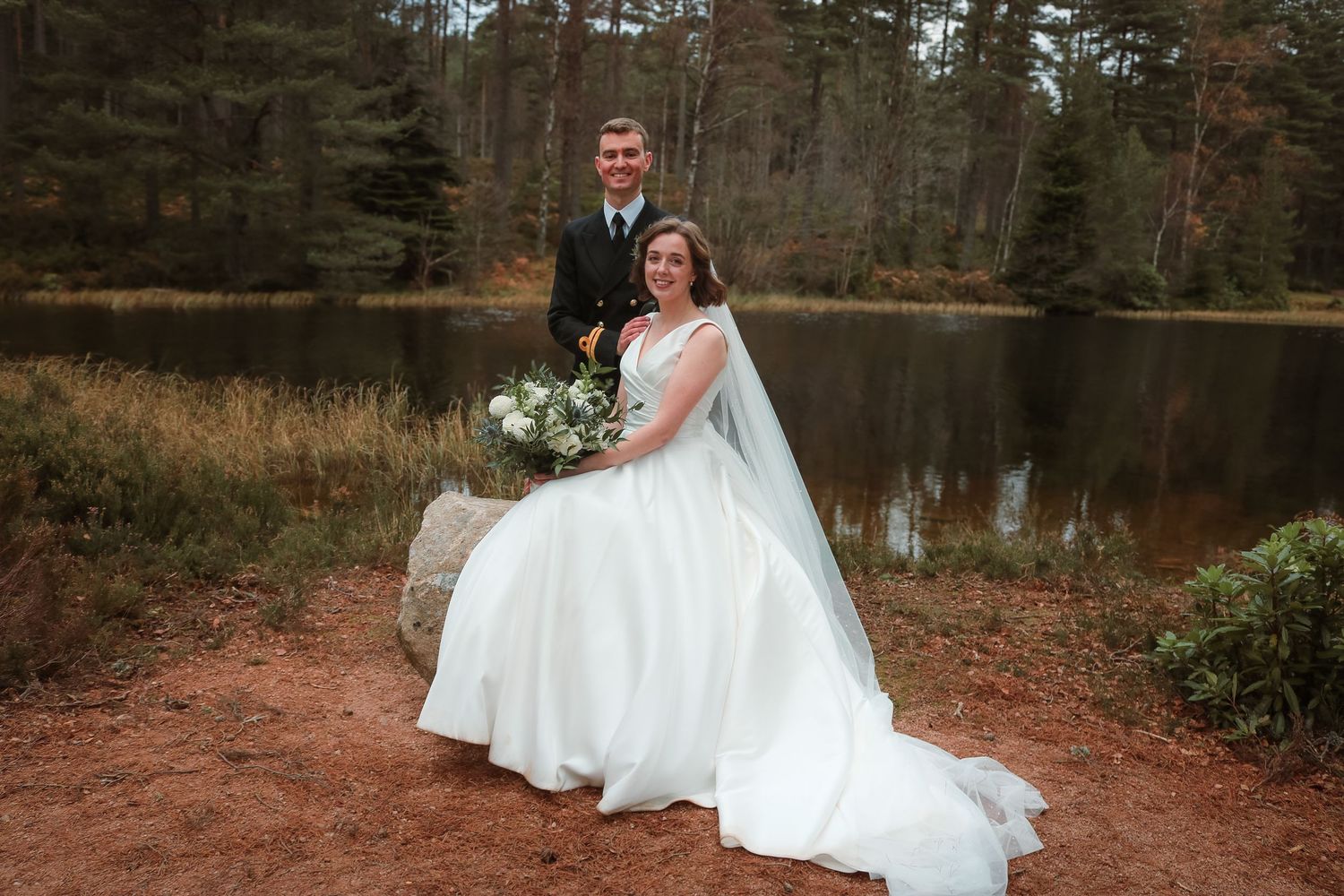 A couple in wedding attire poses beside a tranquil forest lake, with the bride holding a white floral bouquet.