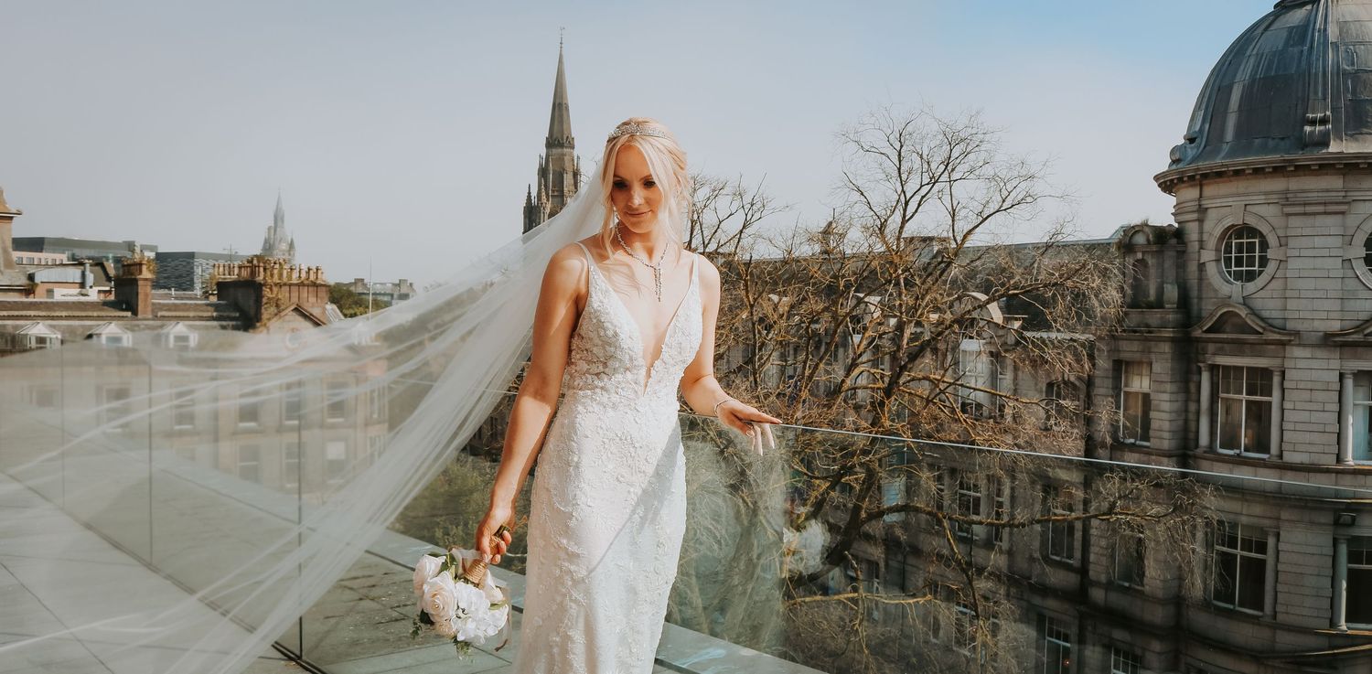 A bride in a sleek white dress poses on a rooftop with historic architecture and church spires in view.