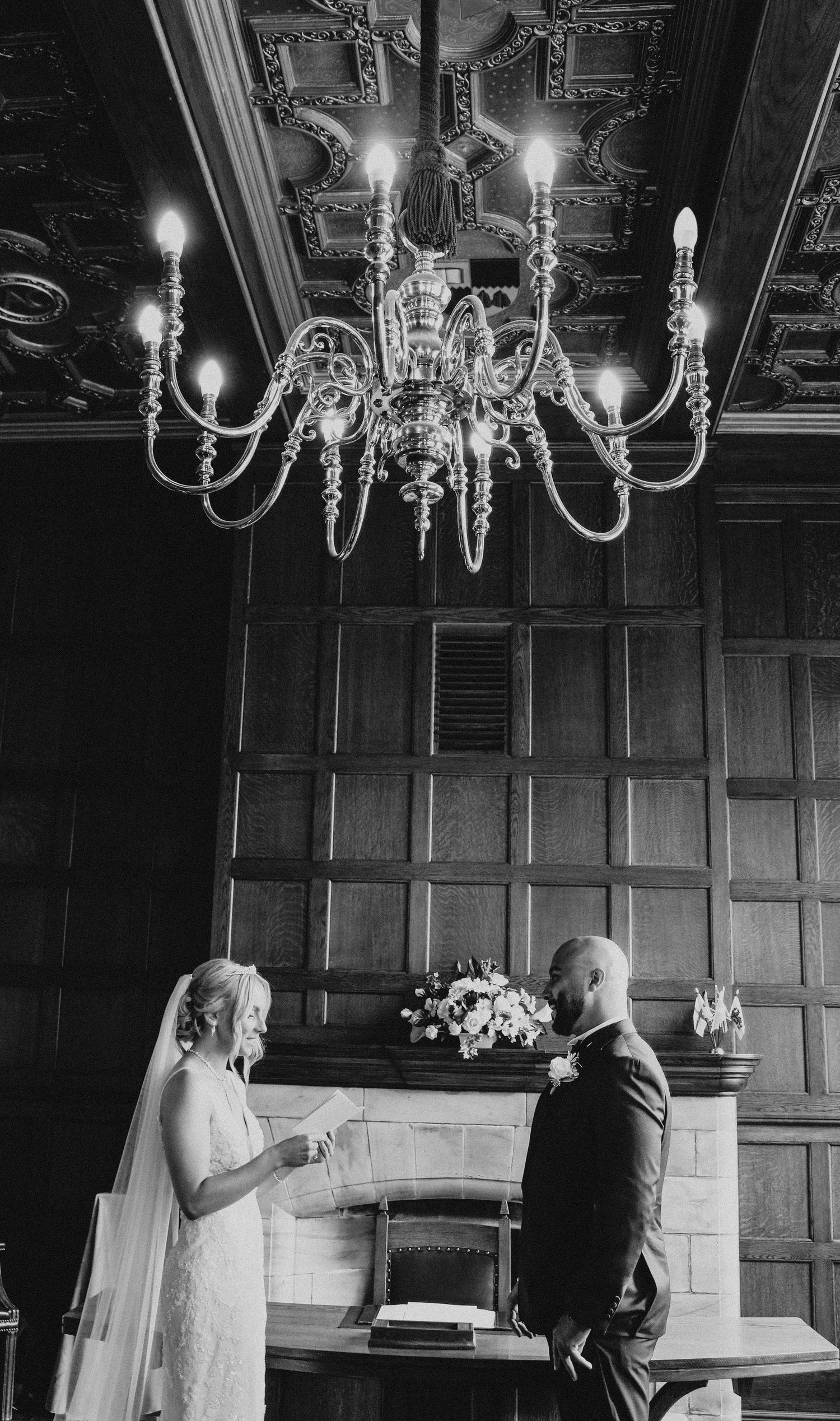 A couple exchanges vows under an elegant chandelier in a wood-paneled room with dramatic lighting.