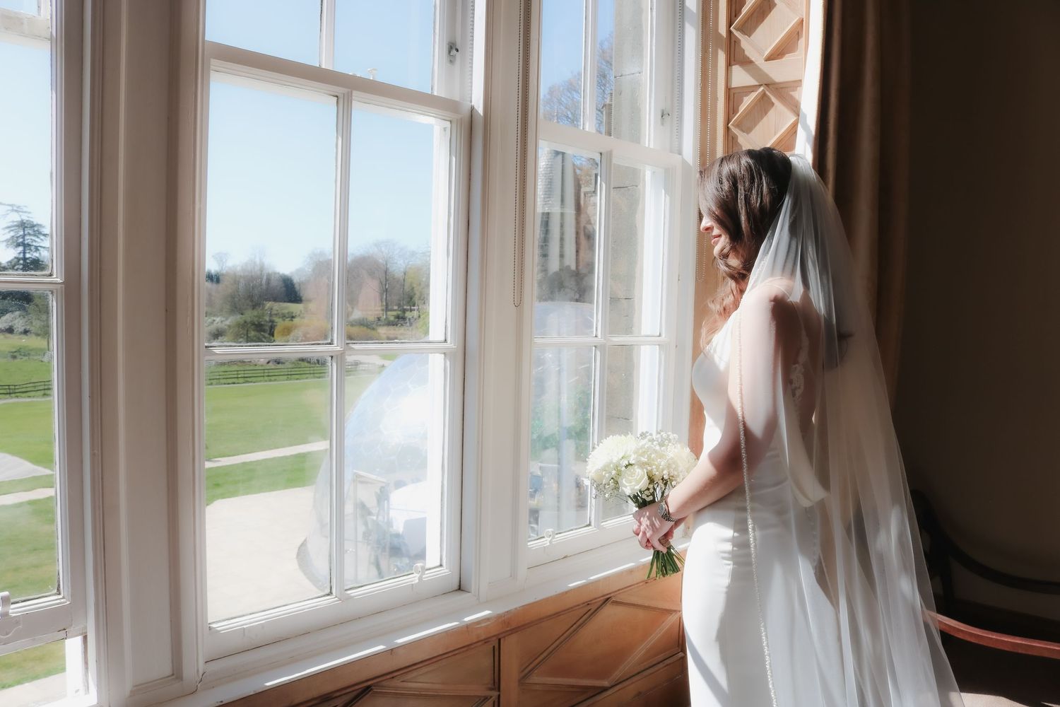 A bride in a white wedding dress and veil gazes out a large window while holding a white floral bouquet.