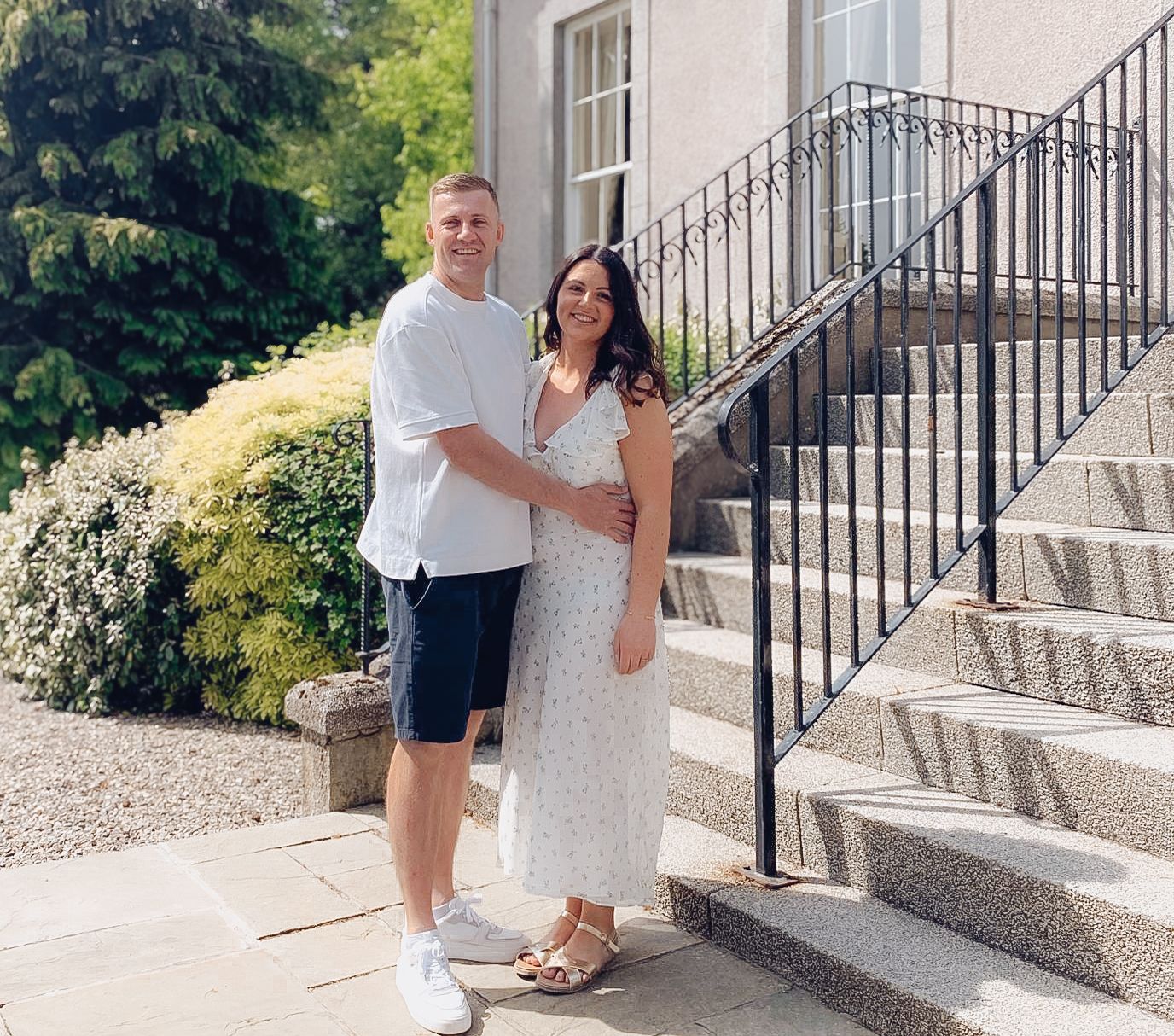 A couple poses together on stone steps with decorative iron railings and landscaping on a sunny summer day.