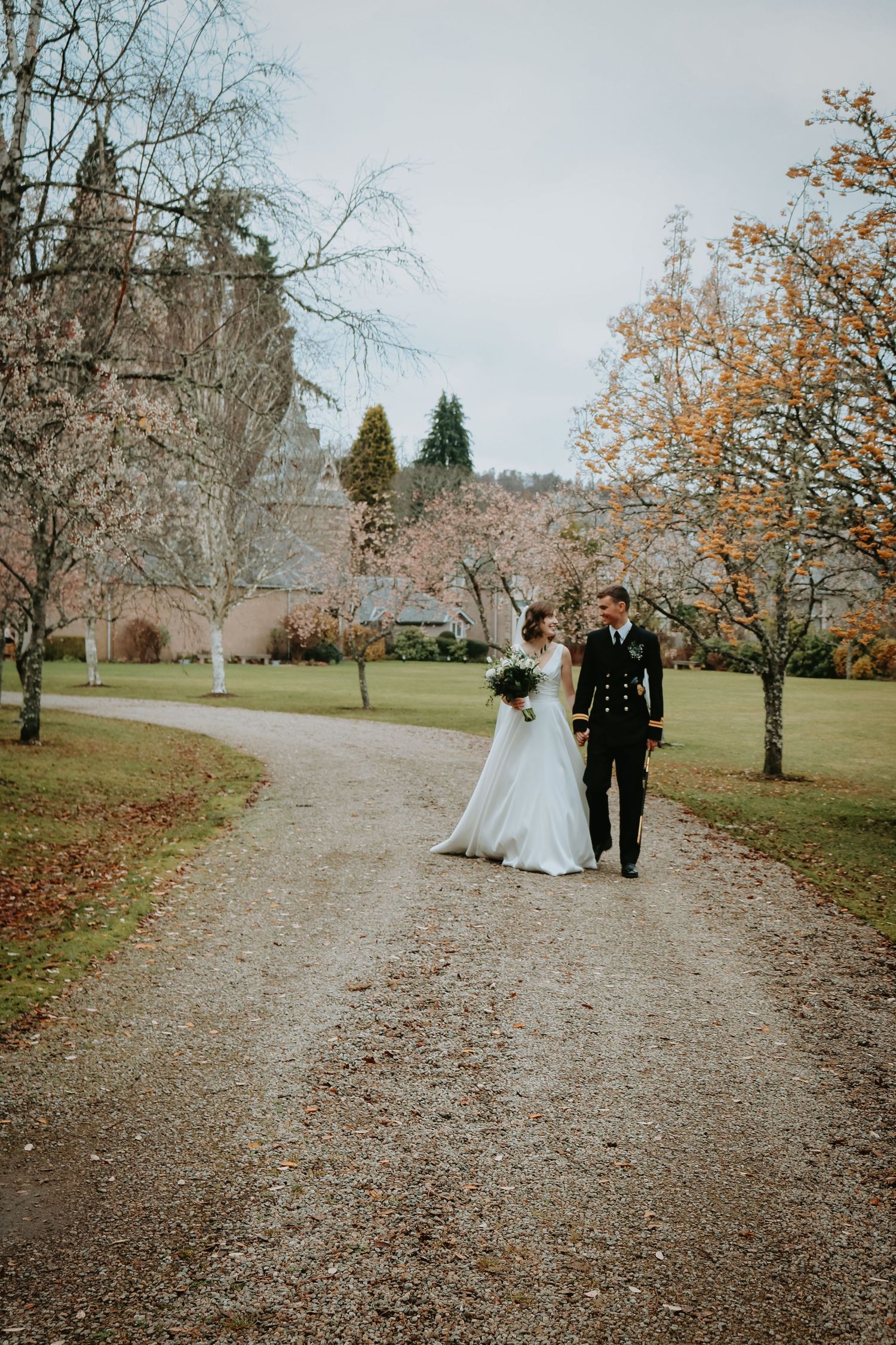 A couple walks down a gravel path surrounded by autumn trees and a historic stone building in the background.