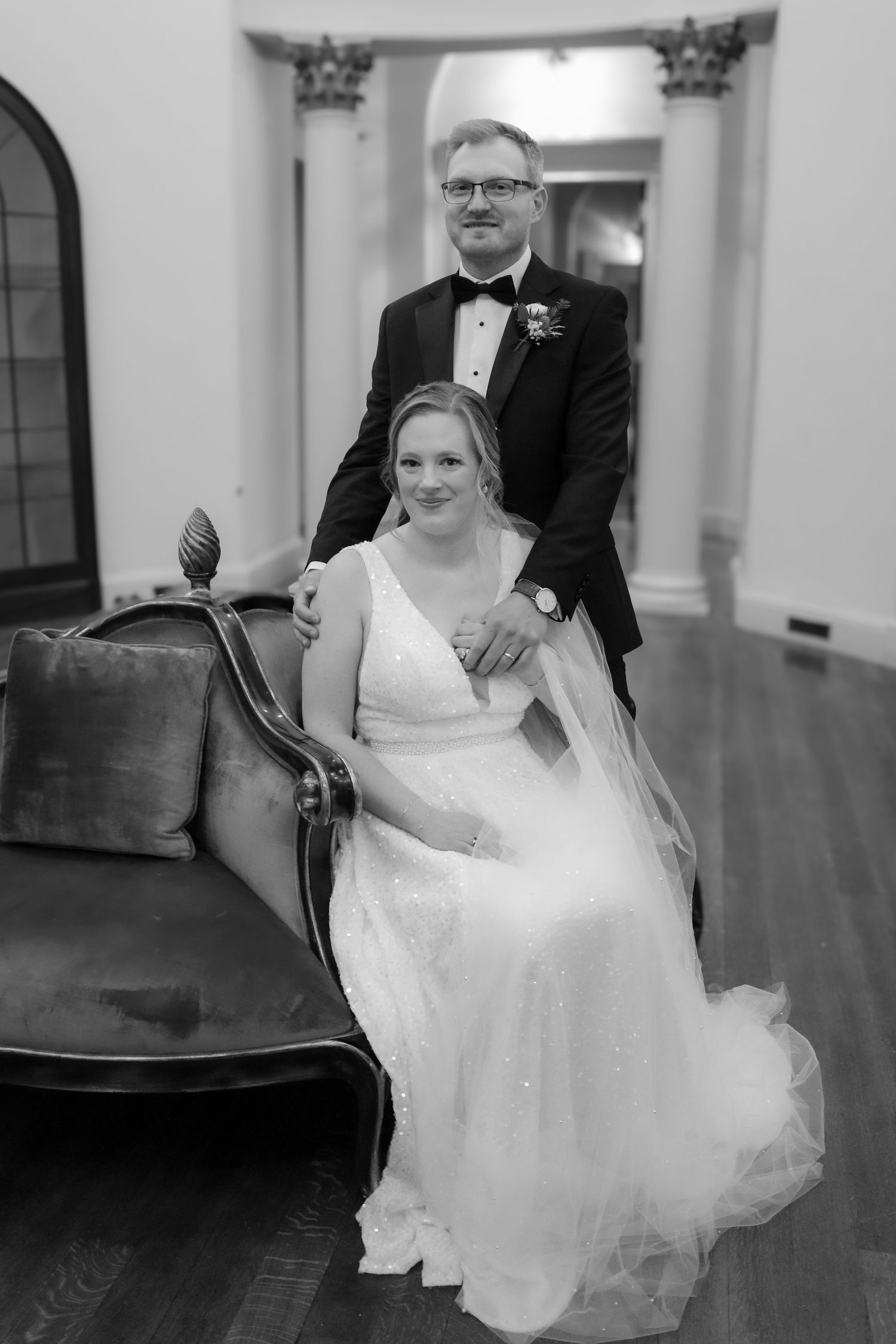 A black and white wedding portrait of newlyweds posing elegantly with bride seated on an antique chair.