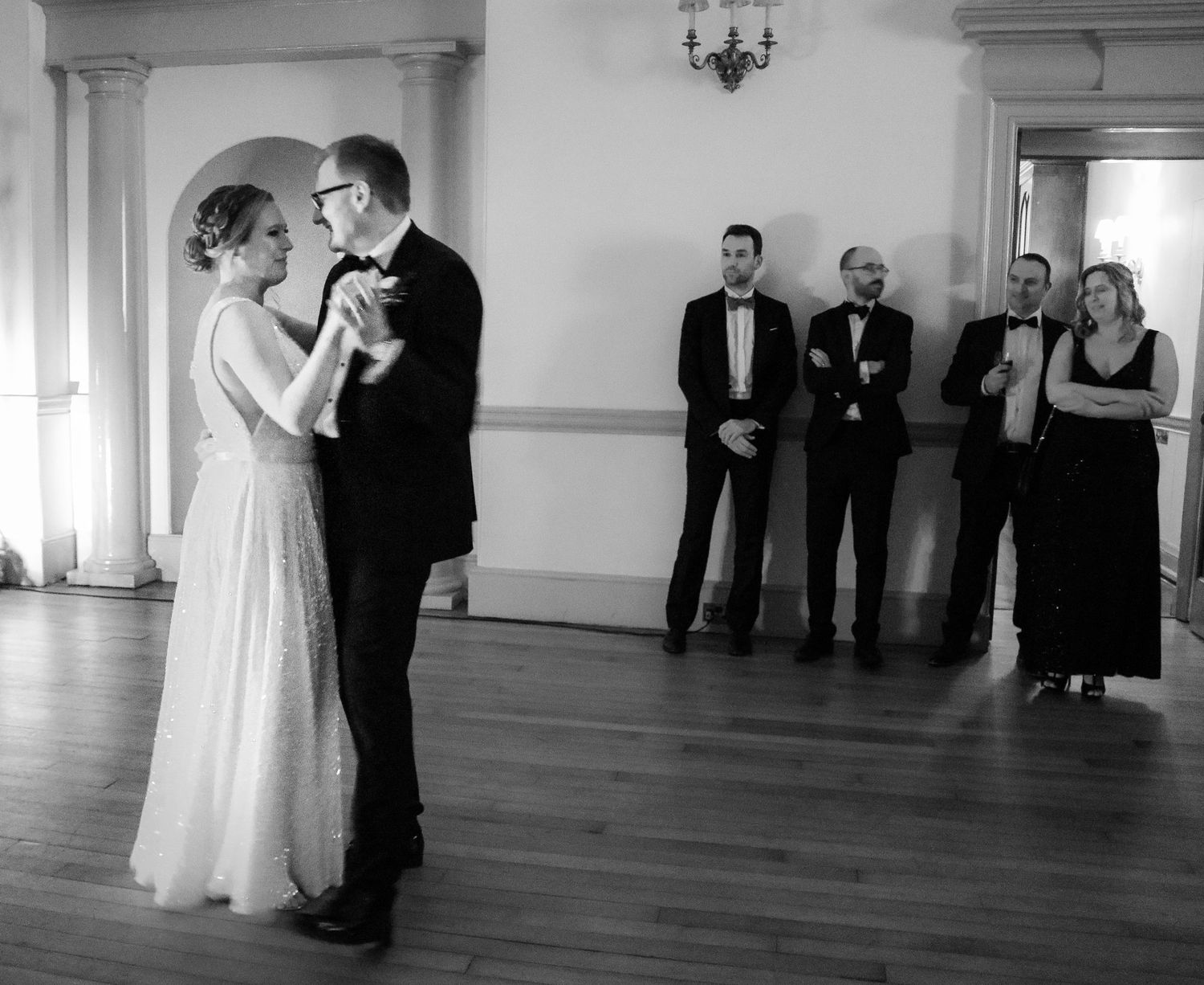 Black and white image of a couple dancing at a wedding reception while guests watch from the side of the room.